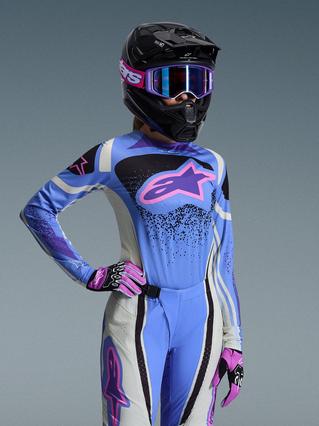 Pilota donna di motocross con indosso il completo Alpinestars 2026 Stella Techstar Nomur, maglia azzurra con accenti rosa e viola, pantaloni coordinati blu e bianchi, casco integrale nero con occhiali viola e guanti rosa e neri, abbigliamento professionale da off-road per donna.