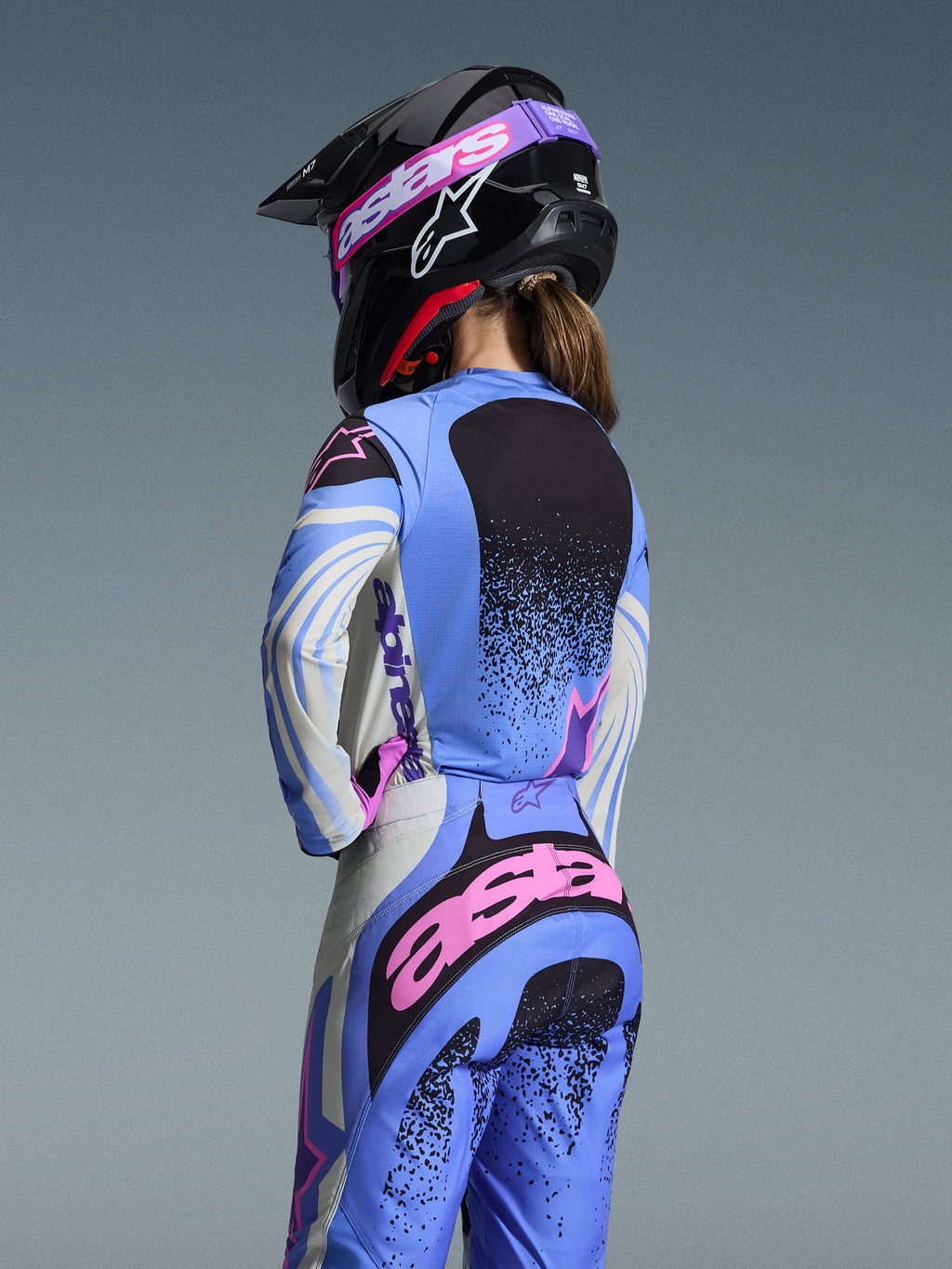 Vista posteriore di una pilota di motocross con il completo Alpinestars 2026 Stella Techstar Nomur, azzurro con accenti rosa e neri, maglia e pantaloni coordinati, casco nero con occhiali rosa, vestibilità ergonomica femminile per gare off-road.