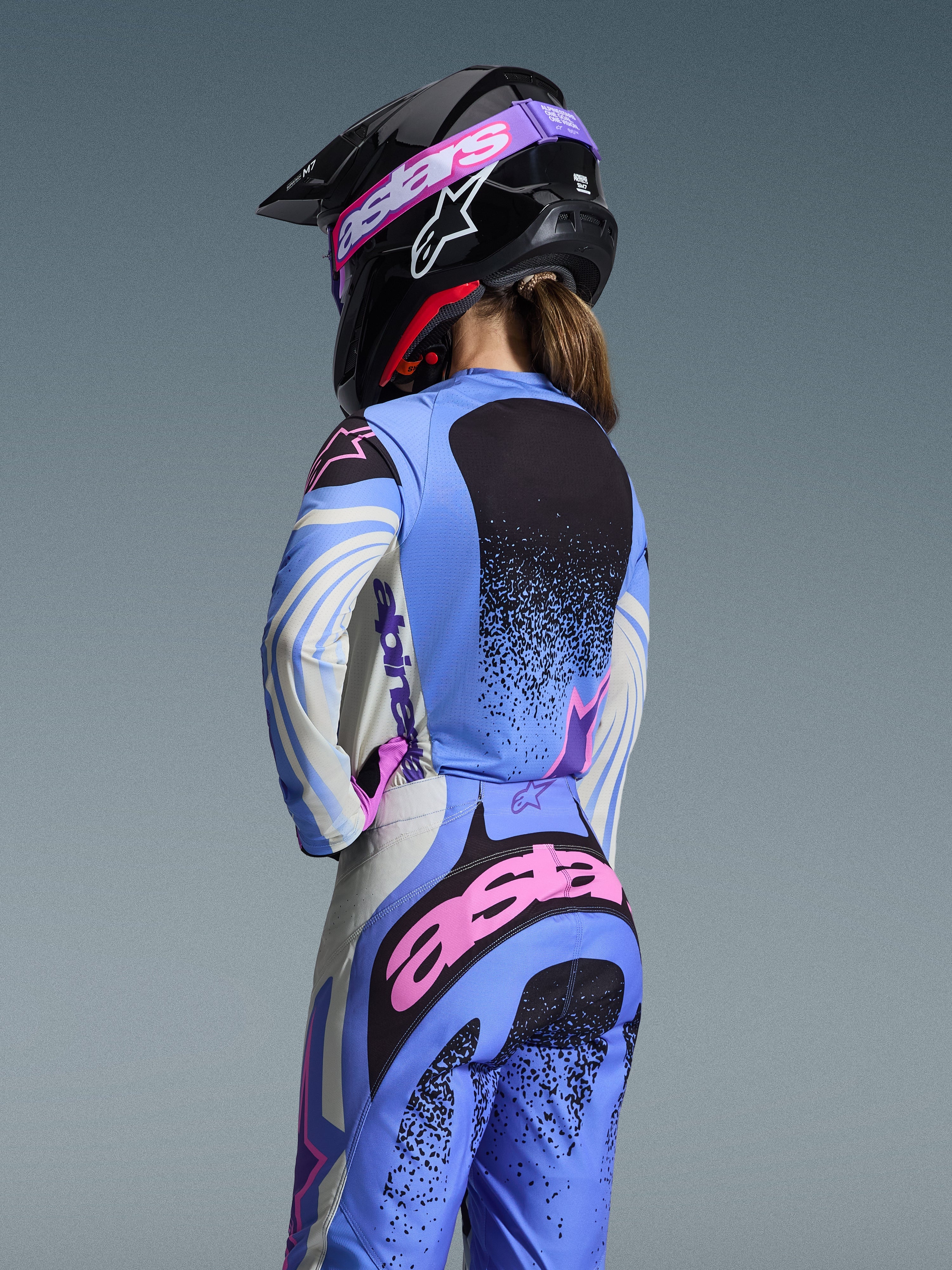 Vista posteriore di una pilota di motocross con il completo Alpinestars 2026 Stella Techstar Nomur, azzurro con accenti rosa e neri, maglia e pantaloni coordinati, casco nero con occhiali rosa, vestibilità ergonomica femminile per gare off-road.