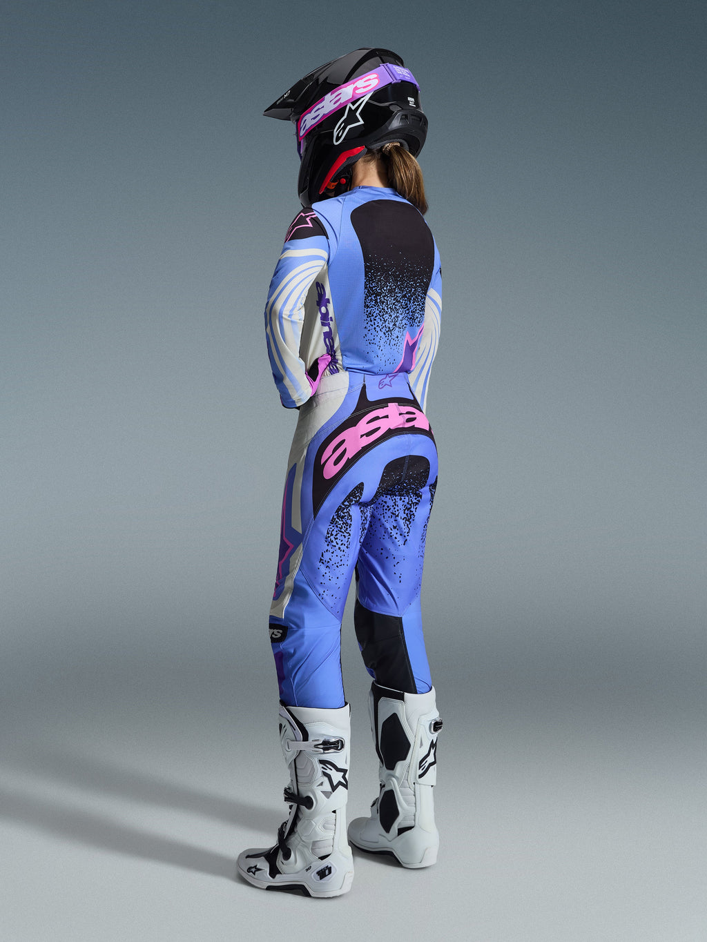 Alpinestars Stella Techstar Nomur Gear Set 2026, vista posteriore di una pilota di motocross con maglia e pantaloni in azzurro, bianco e rosa con dettagli punteggiati neri, casco nero e stivali bianchi, abbigliamento professionale da gara per donna.