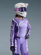 Set completo Alpinestars Stella Fluid Apex 2026, maglia e pantaloni da motocross viola e lavanda, indossati da una pilota con casco bianco e occhiali viola, con loghi e branding Alpinestars bianchi in evidenza