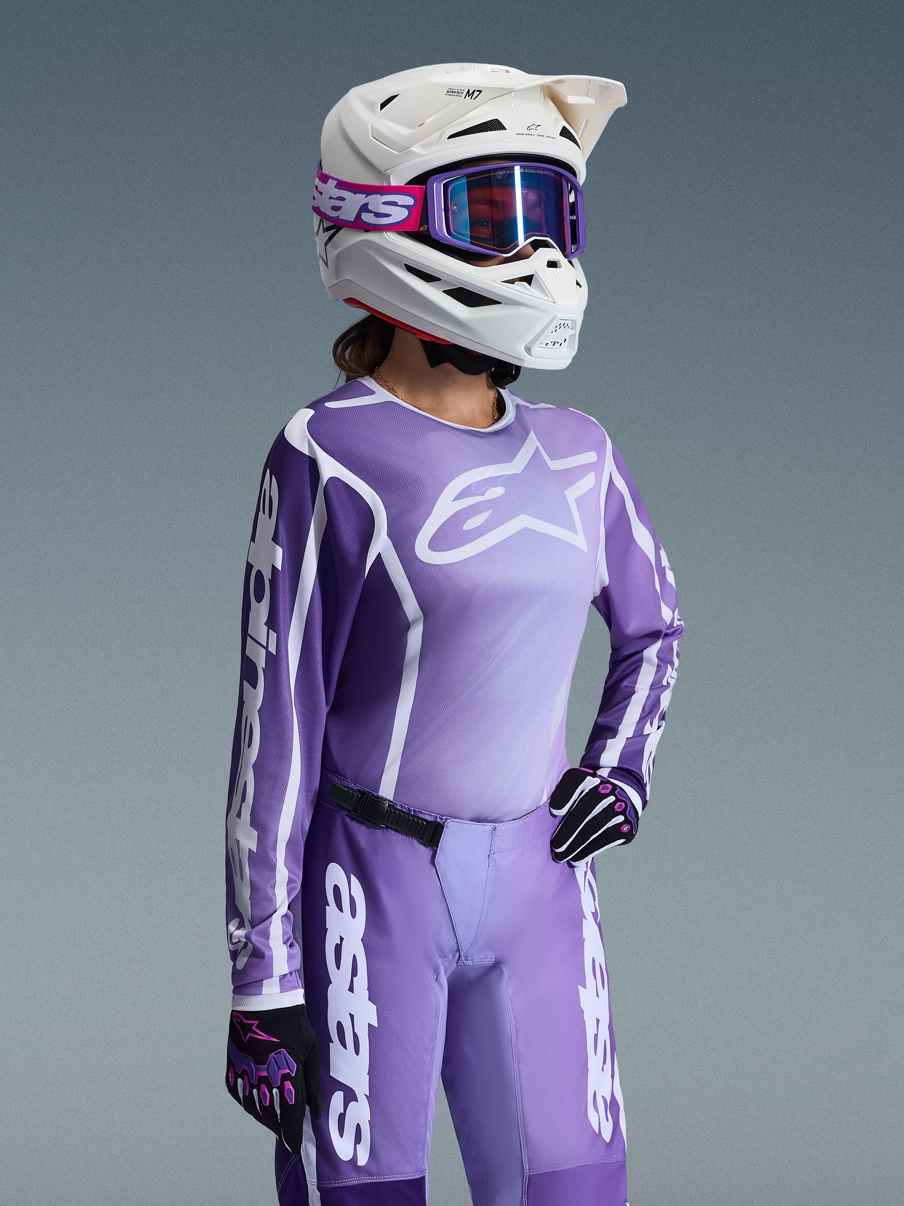 Set completo Alpinestars Stella Fluid Apex 2026, maglia e pantaloni da motocross viola e lavanda, indossati da una pilota con casco bianco e occhiali viola, con loghi e branding Alpinestars bianchi in evidenza