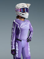 Pilota di motocross con completo Alpinestars Stella Fluid Apex 2026 da donna, maglia viola e bianca con loghi Alpinestars, pantaloni viola coordinati, casco bianco con occhiali viola e guanti neri e viola, abbigliamento professionale da gara off-road