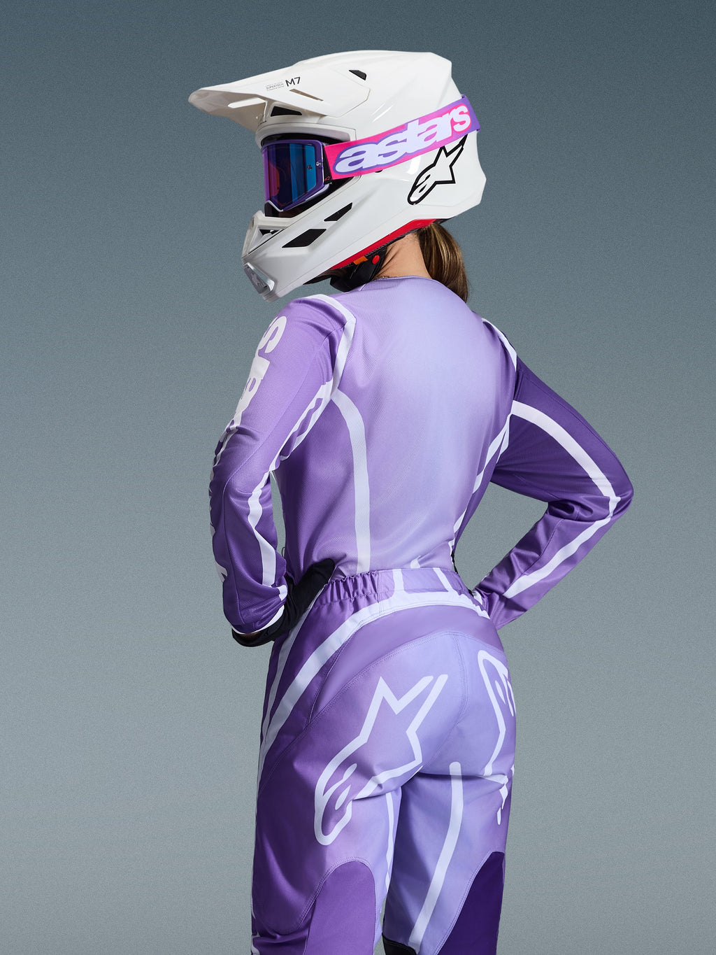 Pilota di motocross con completo Alpinestars Stella Fluid Apex 2026, maglia e pantaloni viola e lavanda con grafiche bianche, casco bianco con occhiali rosa e viola, vestibilità specifica per donna, abbigliamento coordinato per gare off-road