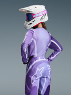 Pilota di motocross con completo Alpinestars Stella Fluid Apex 2026, maglia e pantaloni viola e lavanda con grafiche bianche, casco bianco con occhiali rosa e viola, vestibilità specifica per donna, abbigliamento coordinato per gare off-road