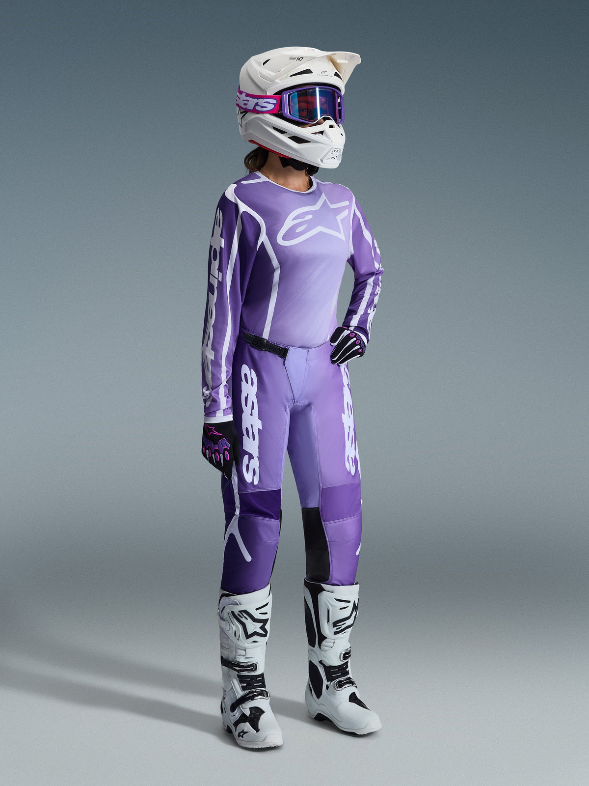 Set completo Alpinestars Stella Fluid Apex 2026, viola e lavanda, pilota di motocross donna che indossa maglia e pantaloni coordinati, casco bianco con occhiali viola, stivali bianchi e neri, guanti neri e viola, abbigliamento completo da performance per corse off-road femminili