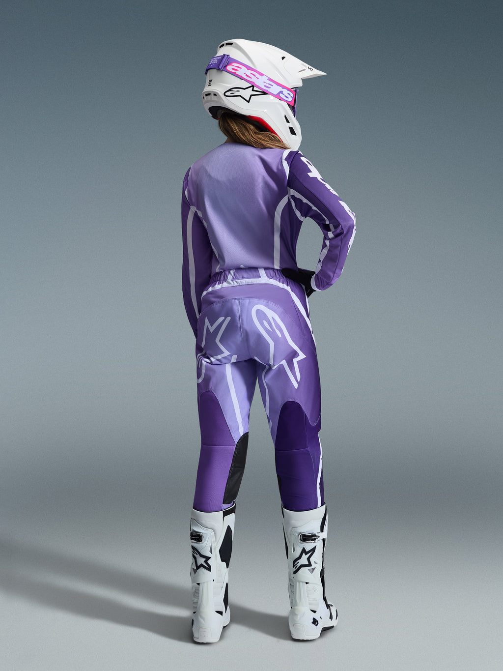 Completo Alpinestars Stella Fluid Apex 2026, abbigliamento da motocross da donna, viola e lavanda con accenti bianchi, pilota donna vista di schiena che indossa maglia e pantaloni coordinati, casco bianco con occhiali rosa e stivali bianchi, outfit da gara coordinato