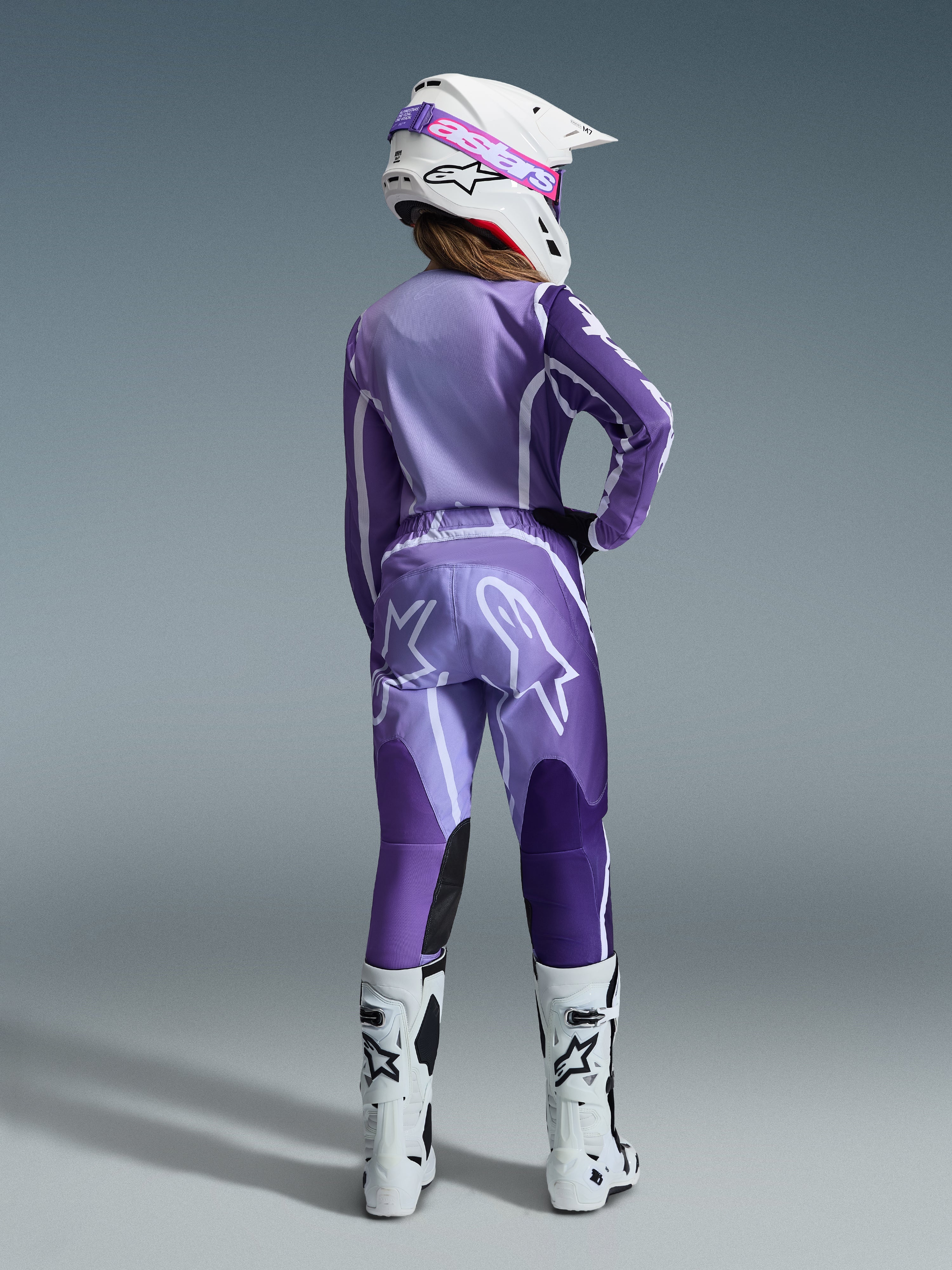 Completo Alpinestars Stella Fluid Apex 2026, abbigliamento da motocross da donna, viola e lavanda con accenti bianchi, pilota donna vista di schiena che indossa maglia e pantaloni coordinati, casco bianco con occhiali rosa e stivali bianchi, outfit da gara coordinato