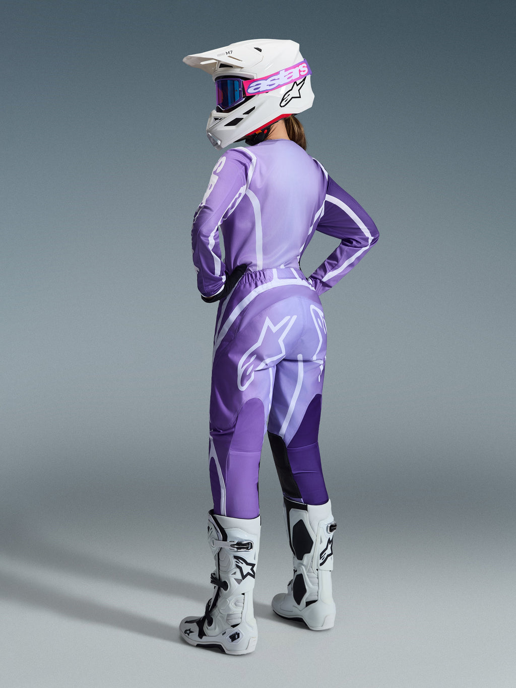 Pilota di motocross donna con completo Stella Fluid Apex 2026, maglia e pantaloni viola e lavanda con dettagli bianchi, casco bianco con maschera rosa, stivali bianchi e neri, abbigliamento coordinato per corse fuoristrada