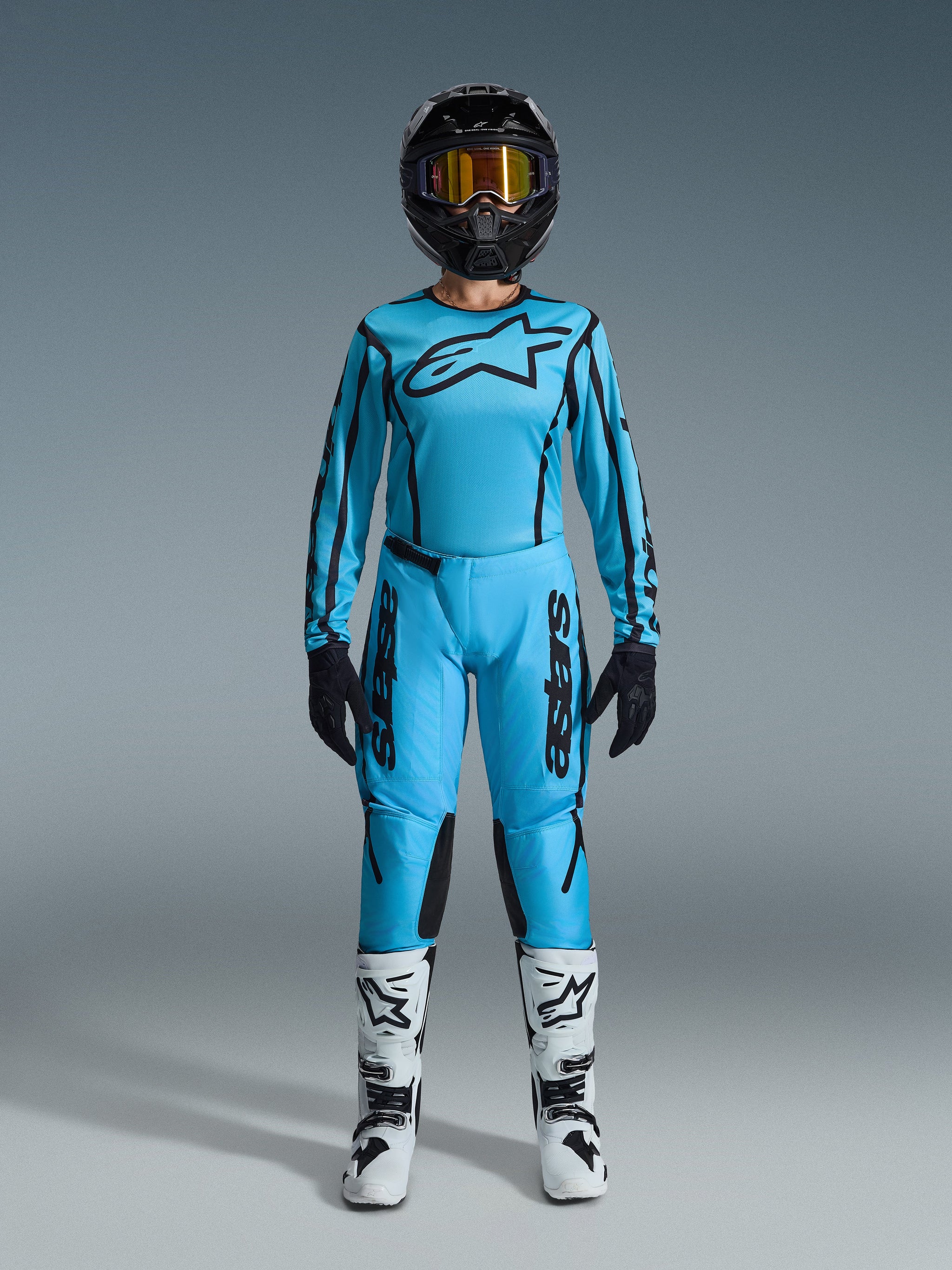 Pilota di motocross con maglia Alpinestars Stella Fluid Apex 2026 e pantaloni coordinati in azzurro con dettagli neri, casco nero con maschera oro, stivali bianchi e neri e guanti neri, set completo di abbigliamento off-road da donna.