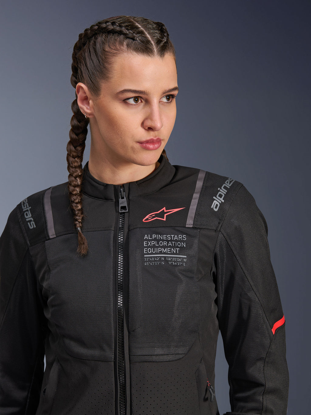 Giacca Alpinestars Stella ST-2 Air, giacca da moto da donna in nero, con pannelli in mesh per il flusso d'aria e marchio 'Alpinestars Exploration Equipment' con coordinate, logo rosso sul petto, indossata da una modella con capelli intrecciati