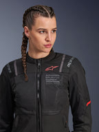 Giacca Alpinestars Stella ST-2 Air, giacca da moto da donna in nero, con pannelli in mesh per il flusso d'aria e marchio 'Alpinestars Exploration Equipment' con coordinate, logo rosso sul petto, indossata da una modella con capelli intrecciati