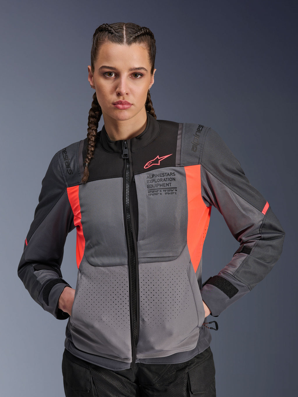 Giacca Alpinestars Stella ST-2 Air, giacca da moto da donna, grigio scuro e corallo fluo, indossata da una modella con capelli intrecciati, caratterizzata da pannelli in mesh tecnica per il flusso d'aria, perforazioni laser e marchio 'Alpinestars Exploration Equipment' sul petto, abbigliamento touring estivo
