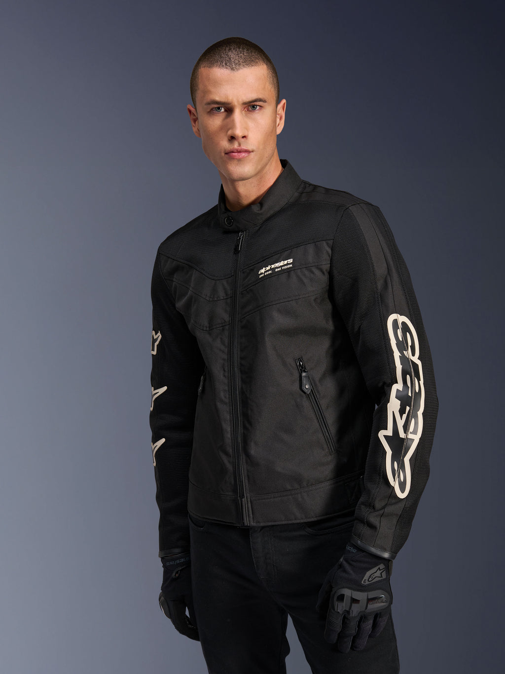 Alpinestars Giacca T-Dyno Air in nero, modello maschile che indossa una giacca da moto in tessuto d'ispirazione vintage con pannelli in mesh per la ventilazione, grande logo Alpinestars bianco sulle maniche, abbinata a guanti da guida neri, design per abbigliamento moto urbano