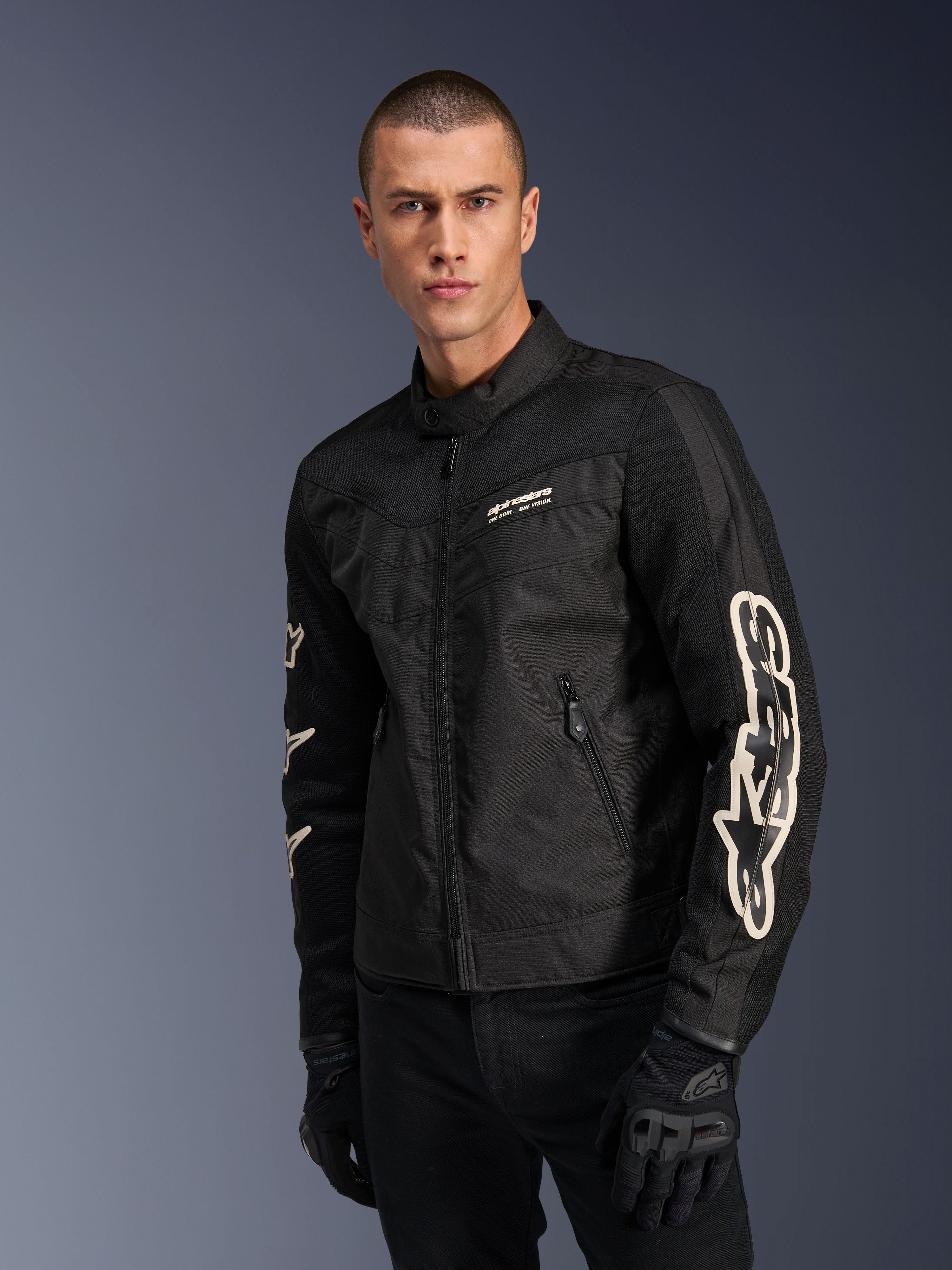 Alpinestars Giacca T-Dyno Air in nero, modello maschile che indossa una giacca da moto in tessuto d'ispirazione vintage con pannelli in mesh per la ventilazione, grande logo Alpinestars bianco sulle maniche, abbinata a guanti da guida neri, design per abbigliamento moto urbano