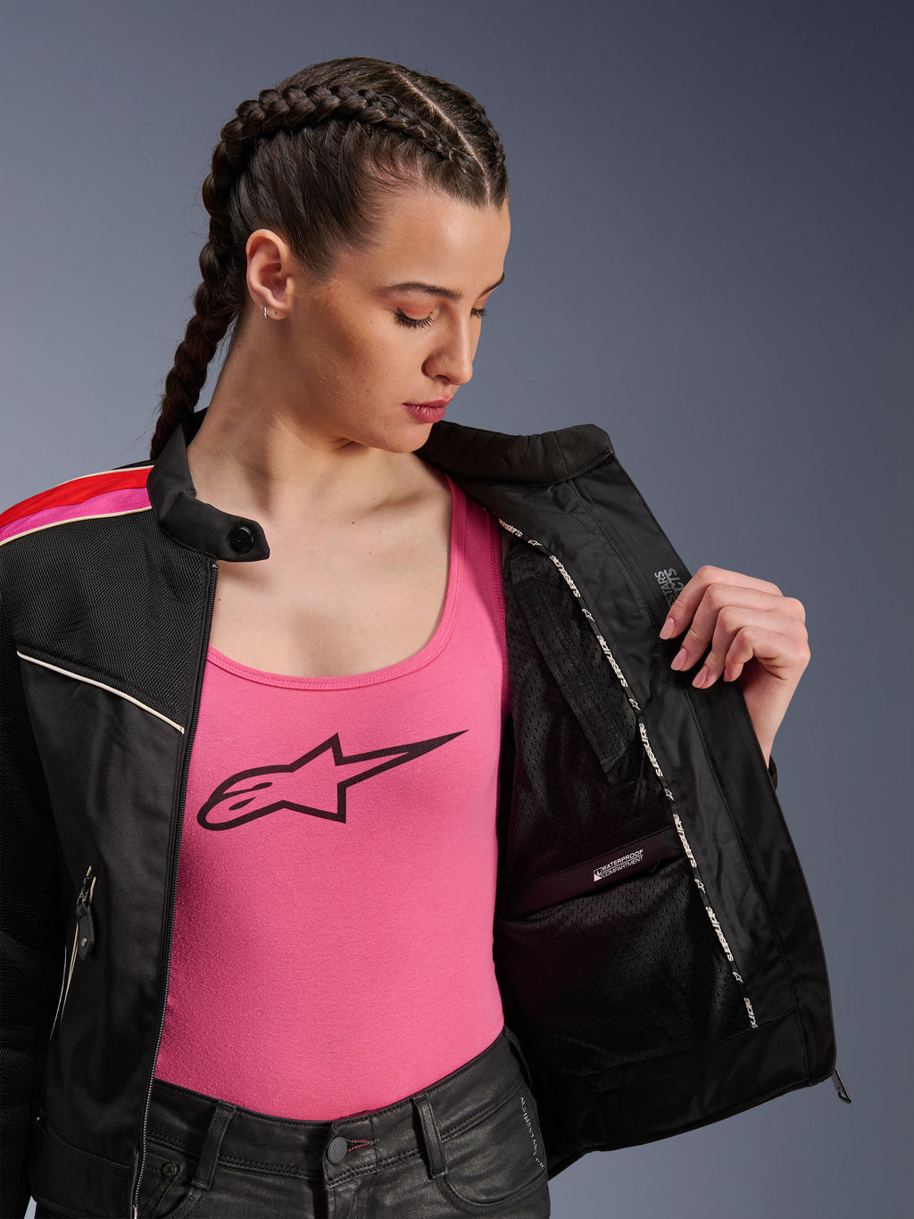 Motociclista che indossa la giacca Alpinestars Stella T-Dyno Air in nero, rosso acceso e fucsia, mostrata parzialmente aperta per rivelare lo scomparto interno impermeabile, indossata sopra una canotta rosa con logo, mettendo in risalto i pannelli in rete traspirante e la vestibilità sartoriale da motociclista.