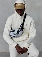Alpinestars Essentials Ageless Hoodie, streetwear casual, Off White, piccolo logo nero ricamato sul petto, cappuccio indossato sopra un berretto color sabbia con marchio ASTARS, abbinata a una borsa a tracolla grigio chiaro, abbigliamento lifestyle premium