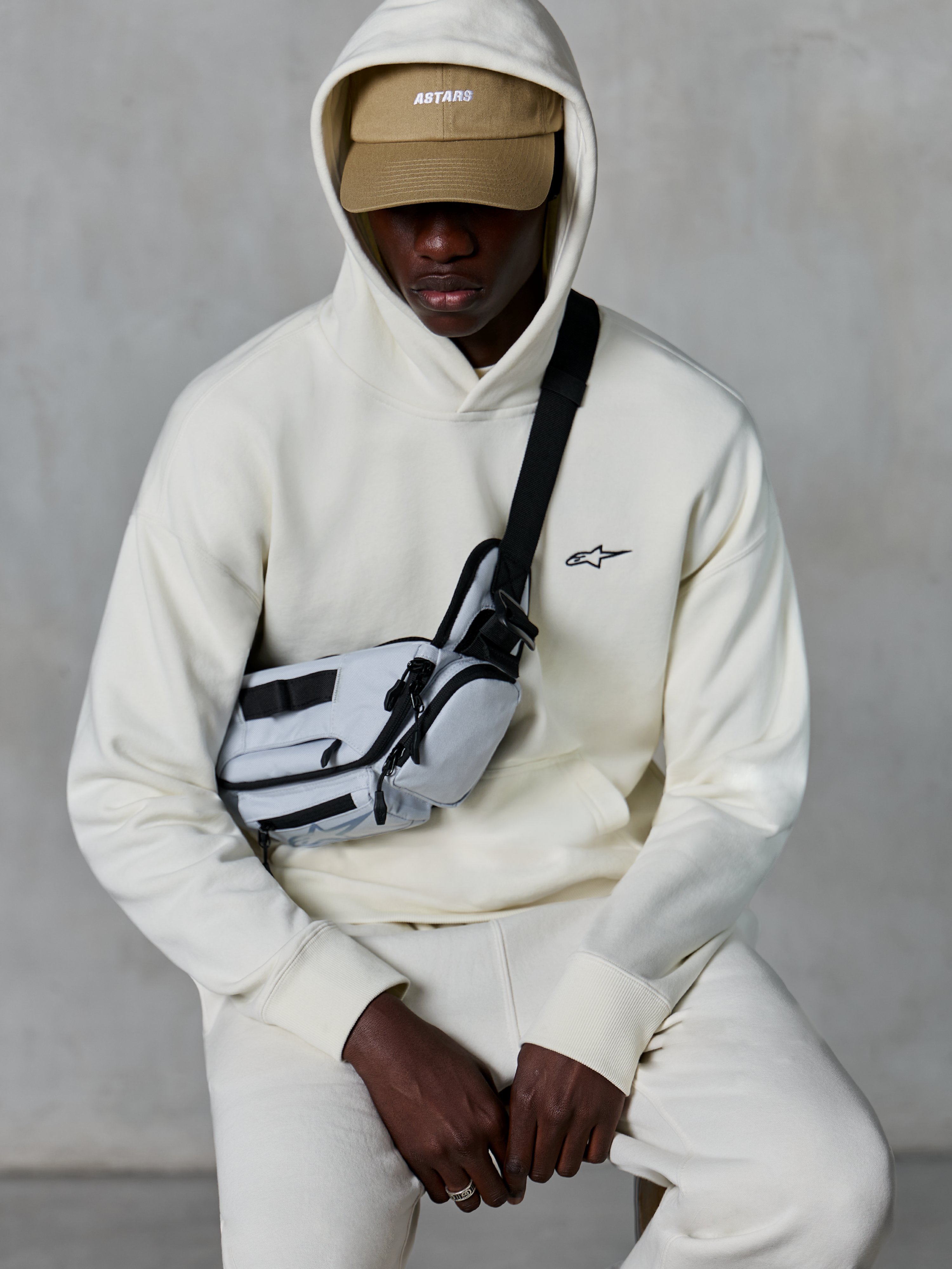 Alpinestars Essentials Ageless Hoodie, streetwear casual, Off White, piccolo logo nero ricamato sul petto, cappuccio indossato sopra un berretto color sabbia con marchio ASTARS, abbinata a una borsa a tracolla grigio chiaro, abbigliamento lifestyle premium