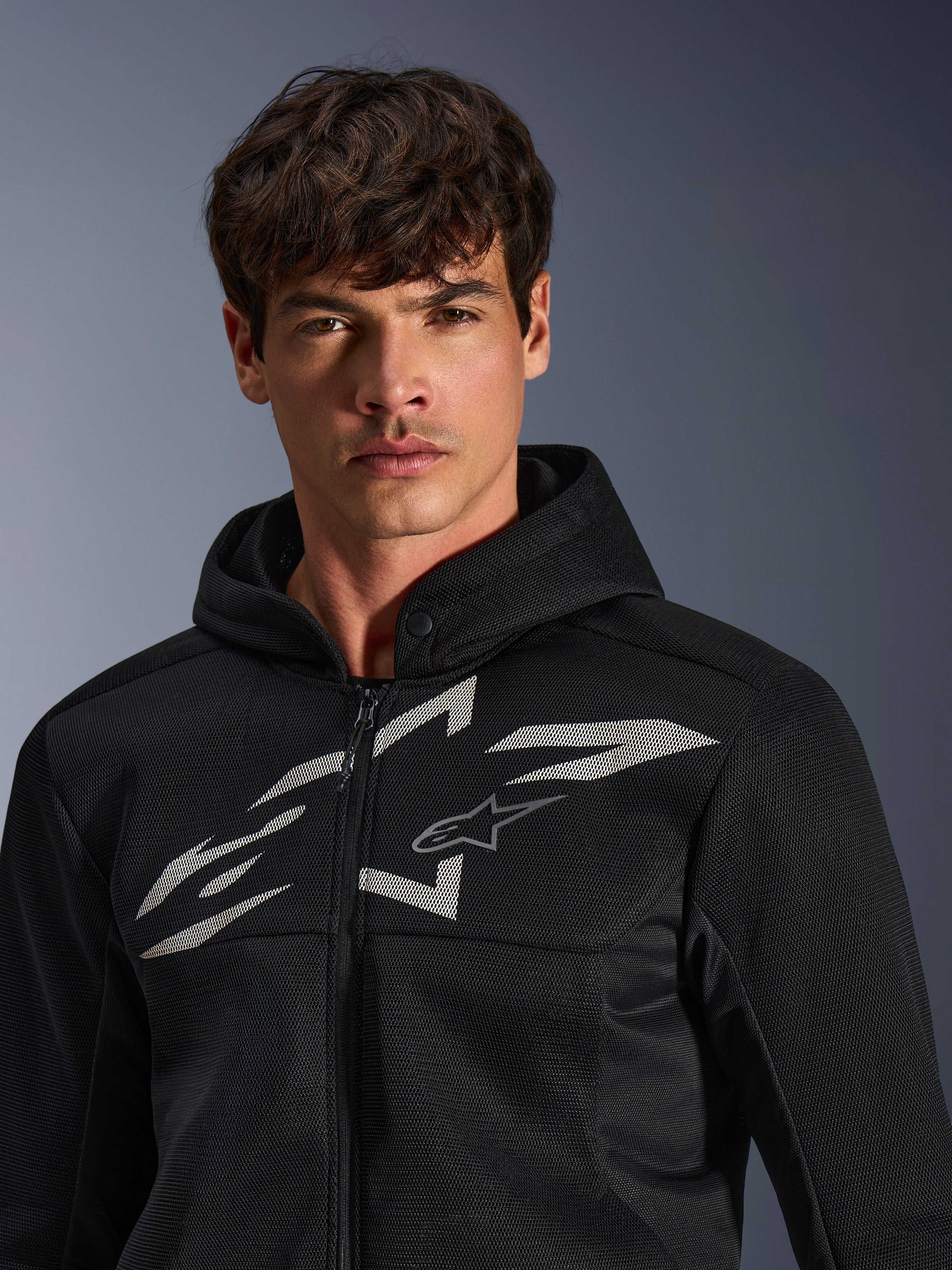 Alpinestars Chrome Superairflow Hoodie, felpa da moto nera, caratterizzata da una costruzione in mesh 3D con un grande logo grafico bianco sul petto, indossata da un modello, abbigliamento per guida sportiva urbana dal moderno design ventilato