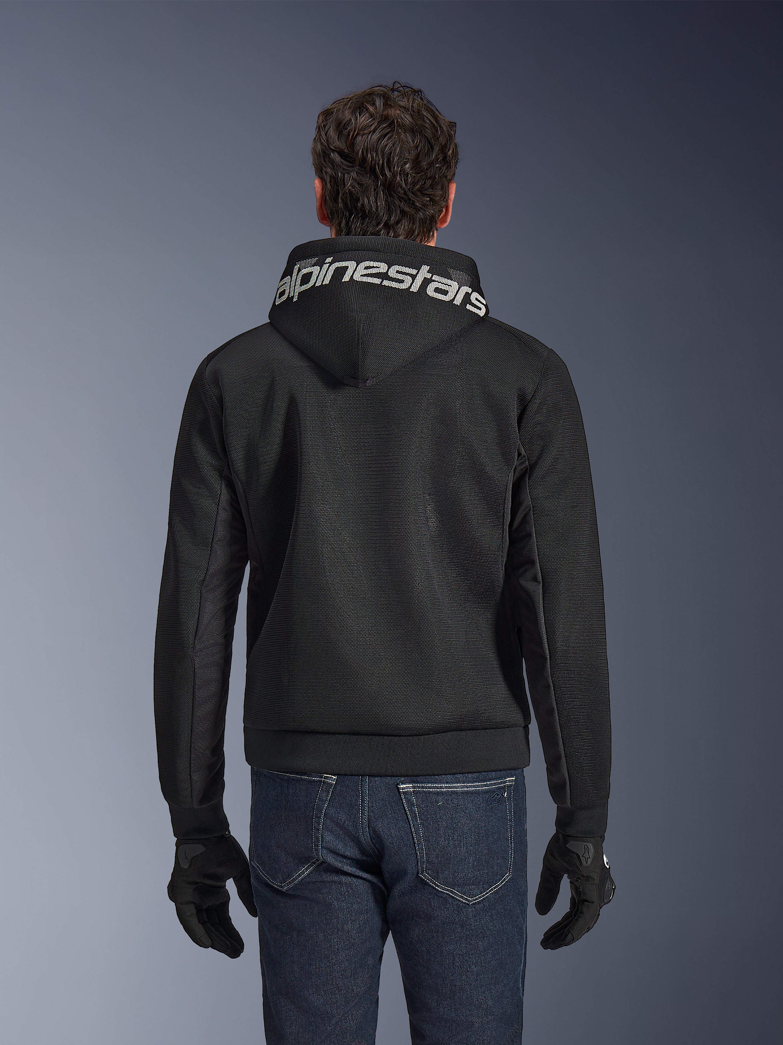 Alpinestars Chrome Superairflow Hoodie, felpa da moto nera, vista posteriore con vistoso marchio Alpinestars bianco sul cappuccio, struttura in mesh tecnico 3D, indossata con jeans blu scuro e guanti neri, abbigliamento da guida urbana ventilato