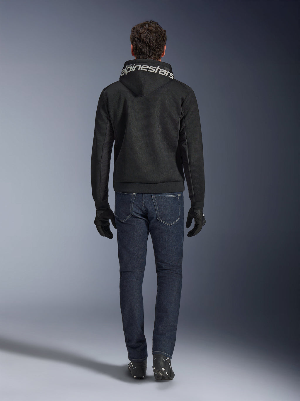 Alpinestars Chrome Superairflow Hoodie, felpa da moto nera con branding bianco sul cappuccio, vista dal retro, indossata con jeans blu scuro, guanti neri e stivali bianchi e neri, abbigliamento urban sport