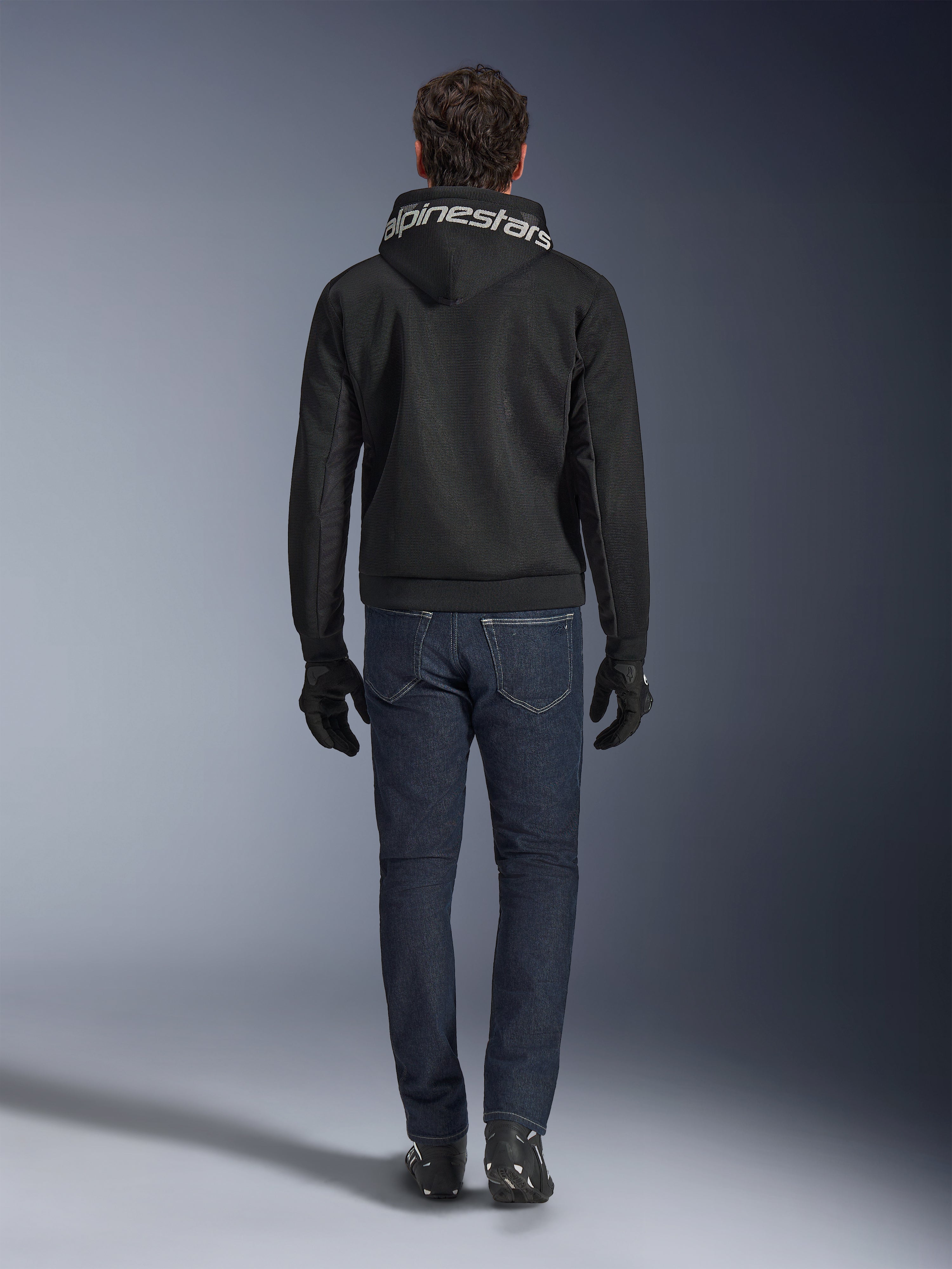Alpinestars Chrome Superairflow Hoodie, felpa da moto nera con branding bianco sul cappuccio, vista dal retro, indossata con jeans blu scuro, guanti neri e stivali bianchi e neri, abbigliamento urban sport