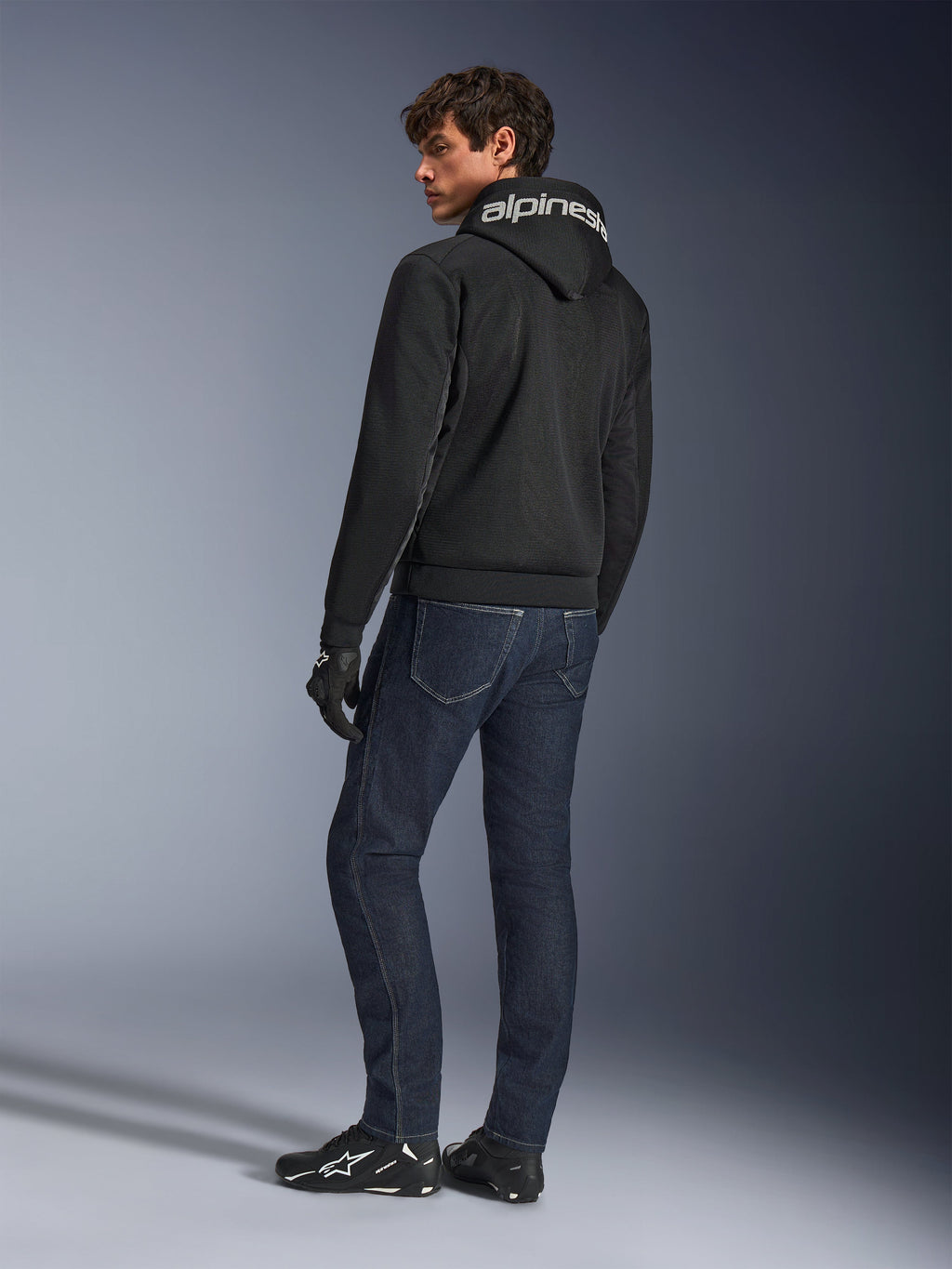 Alpinestars Chrome Superairflow Hoodie, nero, vista posteriore su modello, con marchio Alpinestars bianco sul cappuccio, indossata con jeans in denim scuro e guanti neri, design per abbigliamento moto urbano