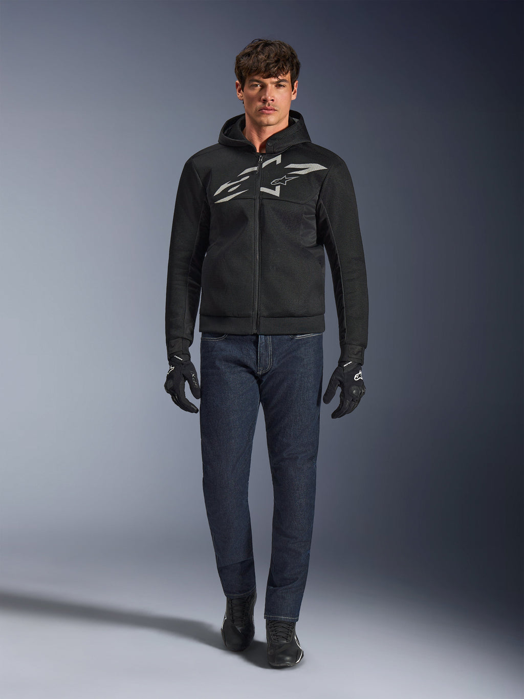Motociclista che indossa la felpa Alpinestars Chrome Superairflow in nero e grigio, abbinata a jeans blu scuro, guanti da moto neri e scarpe nere. La felpa presenta un grande logo stilizzato sul petto e una trama in mesh 3D per prestazioni elevate con alte temperature.