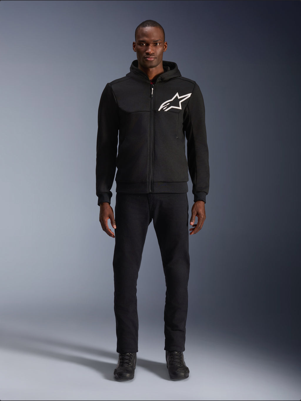 Alpinestars Chrome V2 Sport Hoodie, felpa da moto nera e bianca, grande logo Alpinestars bianco sul petto, zip frontale e cappuccio, indossata da un modello con pantaloni e stivali neri, abbigliamento protettivo da guida urbana