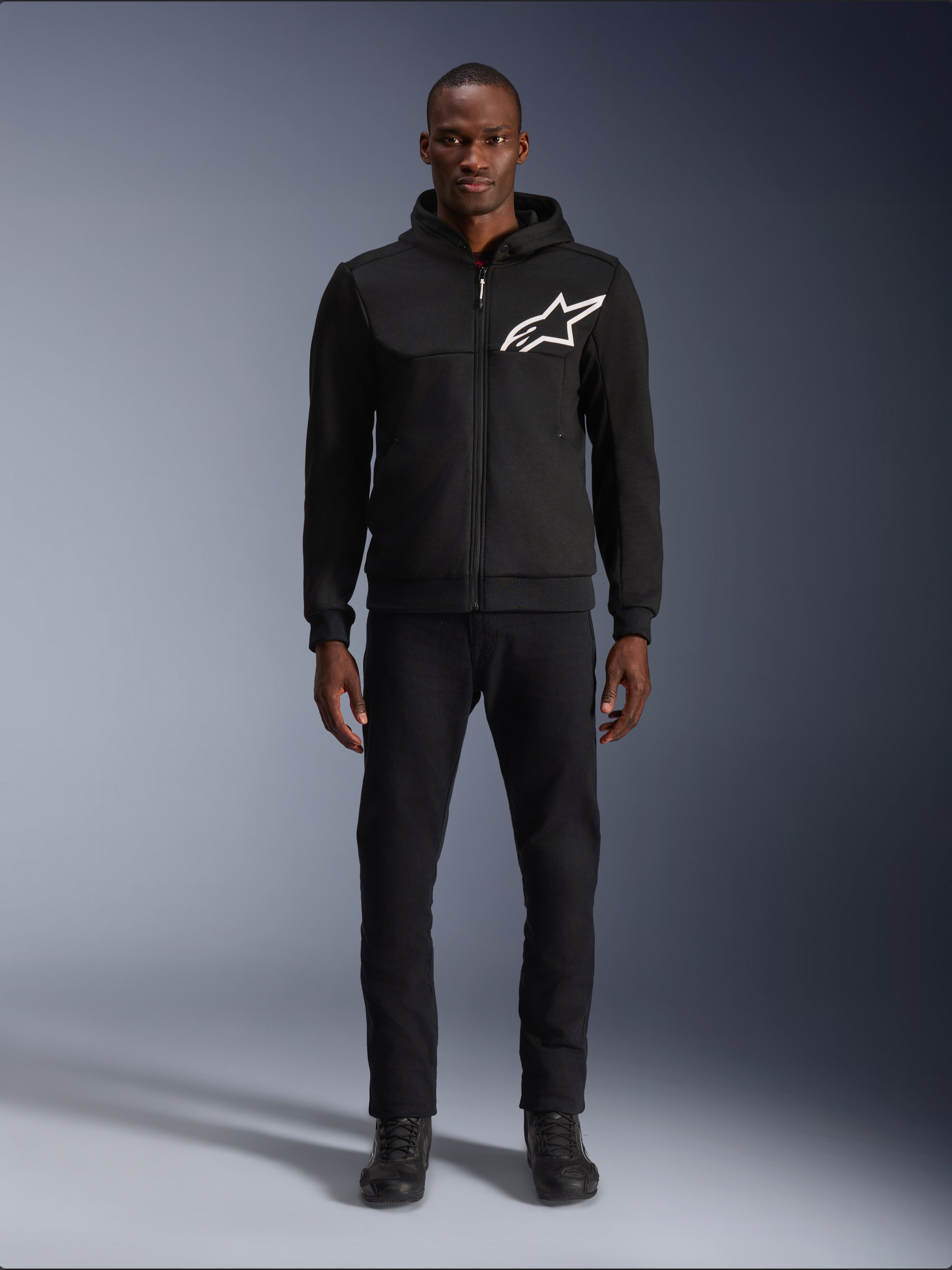 Alpinestars Chrome V2 Sport Hoodie, felpa da moto nera e bianca, grande logo Alpinestars bianco sul petto, zip frontale e cappuccio, indossata da un modello con pantaloni e stivali neri, abbigliamento protettivo da guida urbana