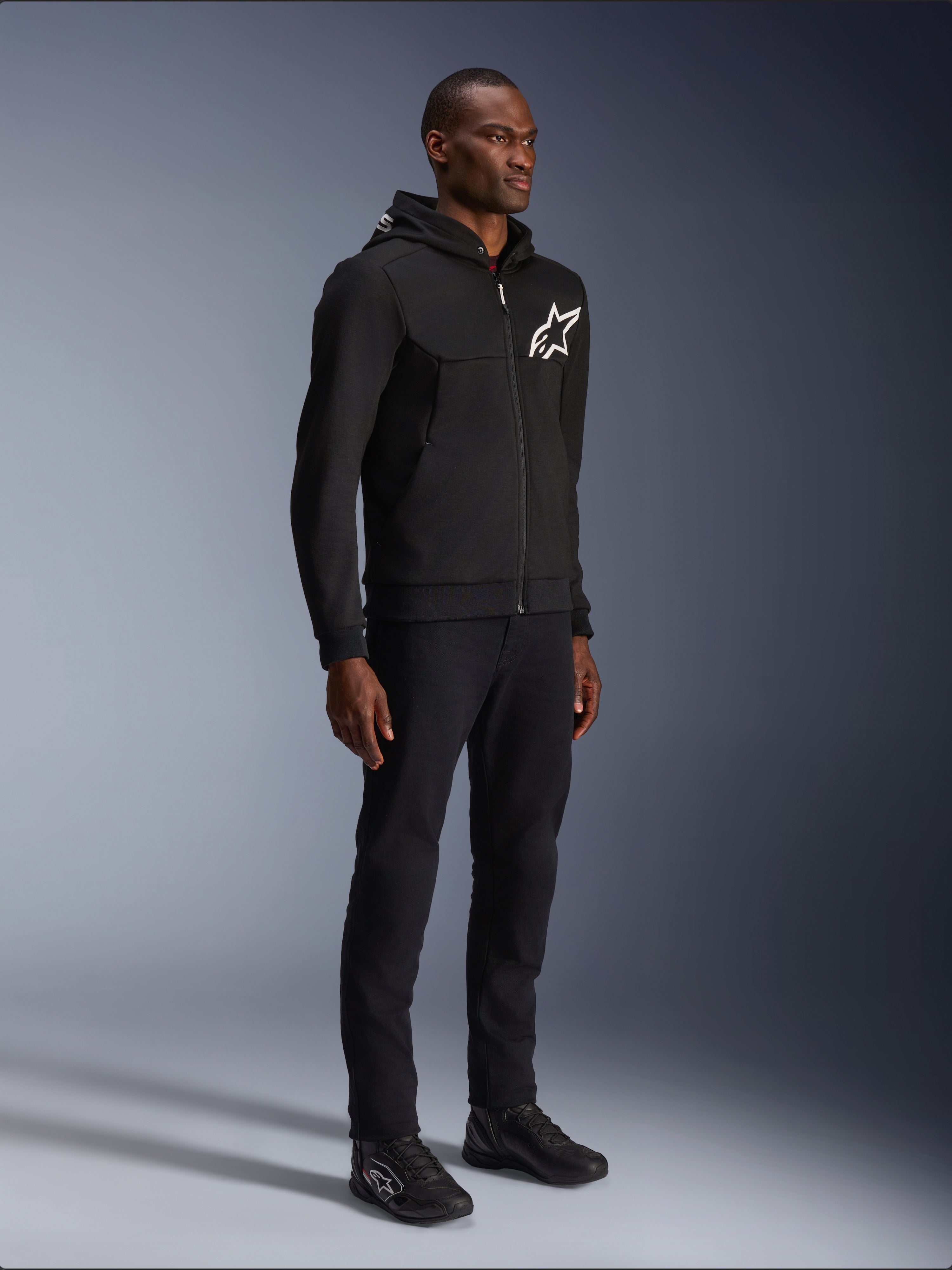 Alpinestars Chrome V2 Sport Hoodie, felpa da moto nera con logo a stella bianco, indossata da un modello con pantaloni neri coordinati e scarpe da moto, abbigliamento da guida urban lifestyle