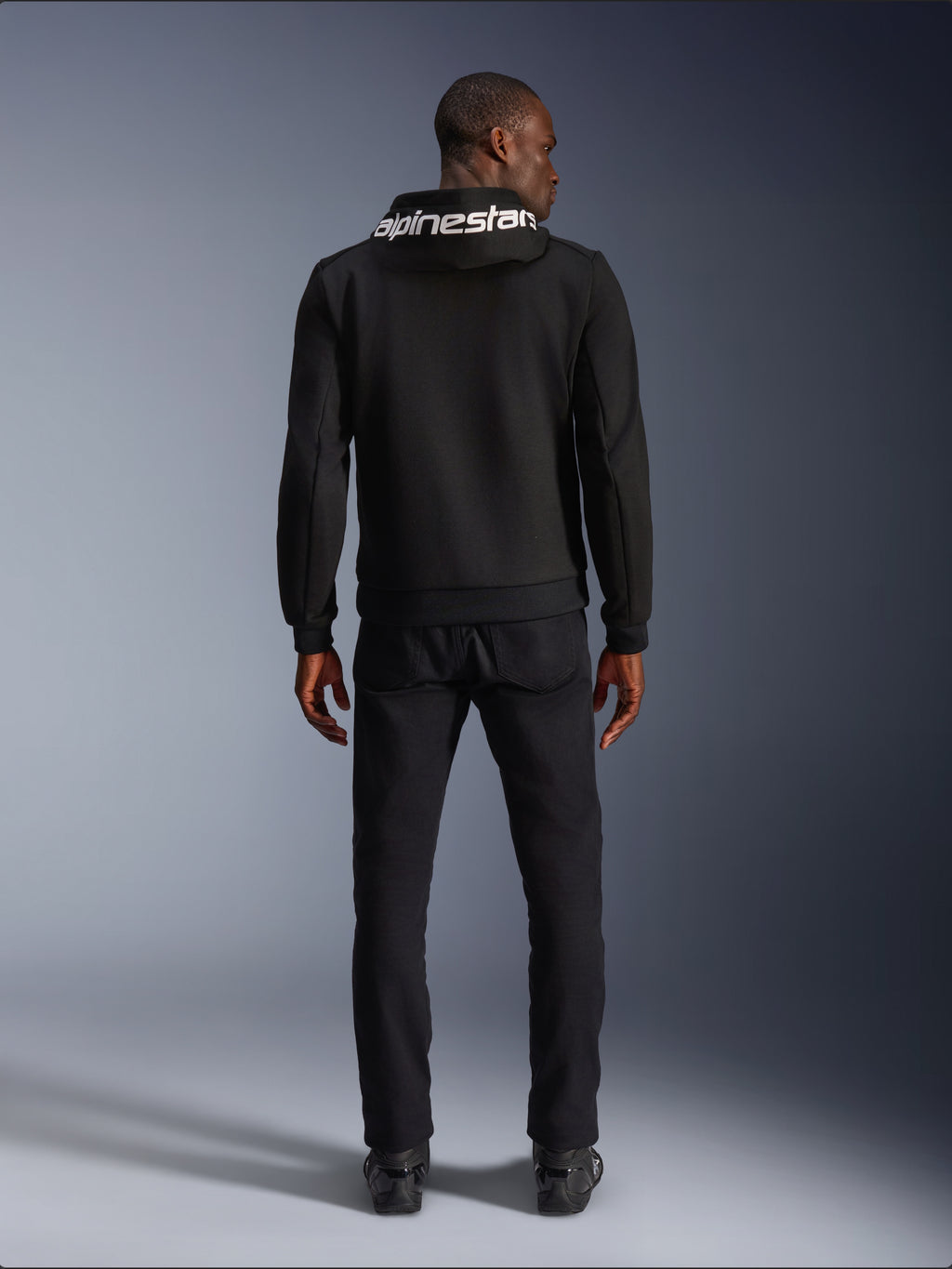 Alpinestars Chrome V2 Sport Hoodie, felpa da moto, nero, vista posteriore del modello che indossa una felpa in pile tecnico nera con logo Alpinestars bianco sul cappuccio, abbinata a pantaloni e stivali neri, stile urban riding casual