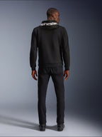Alpinestars Chrome V2 Sport Hoodie, felpa da moto, nero, vista posteriore del modello che indossa una felpa in pile tecnico nera con logo Alpinestars bianco sul cappuccio, abbinata a pantaloni e stivali neri, stile urban riding casual