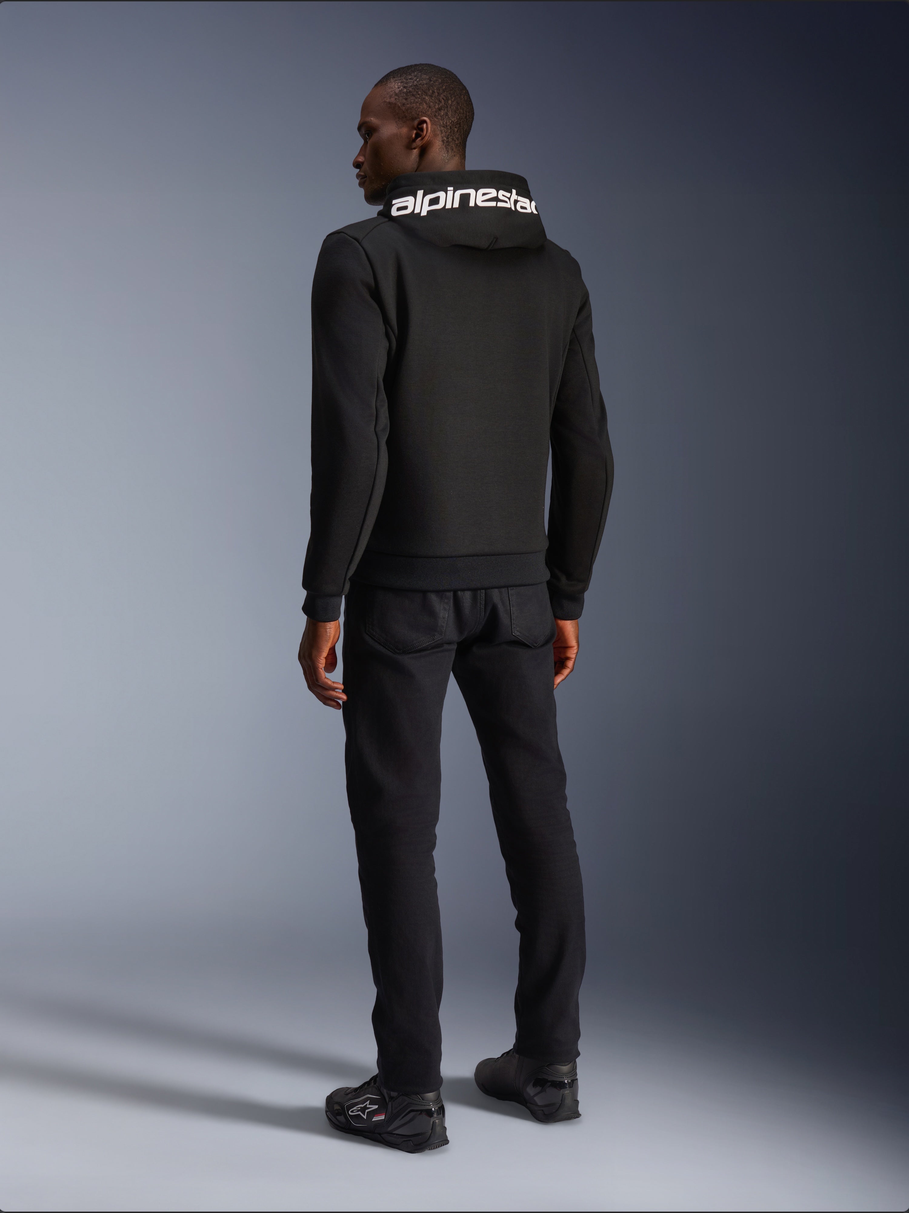 Alpinestars Chrome V2 Sport Hoodie, nero, vista posteriore con branding Alpinestars bianco sul cappuccio, indossata con pantaloni neri e scarpe da moto, abbigliamento da moto urban casual dalla silhouette atletica