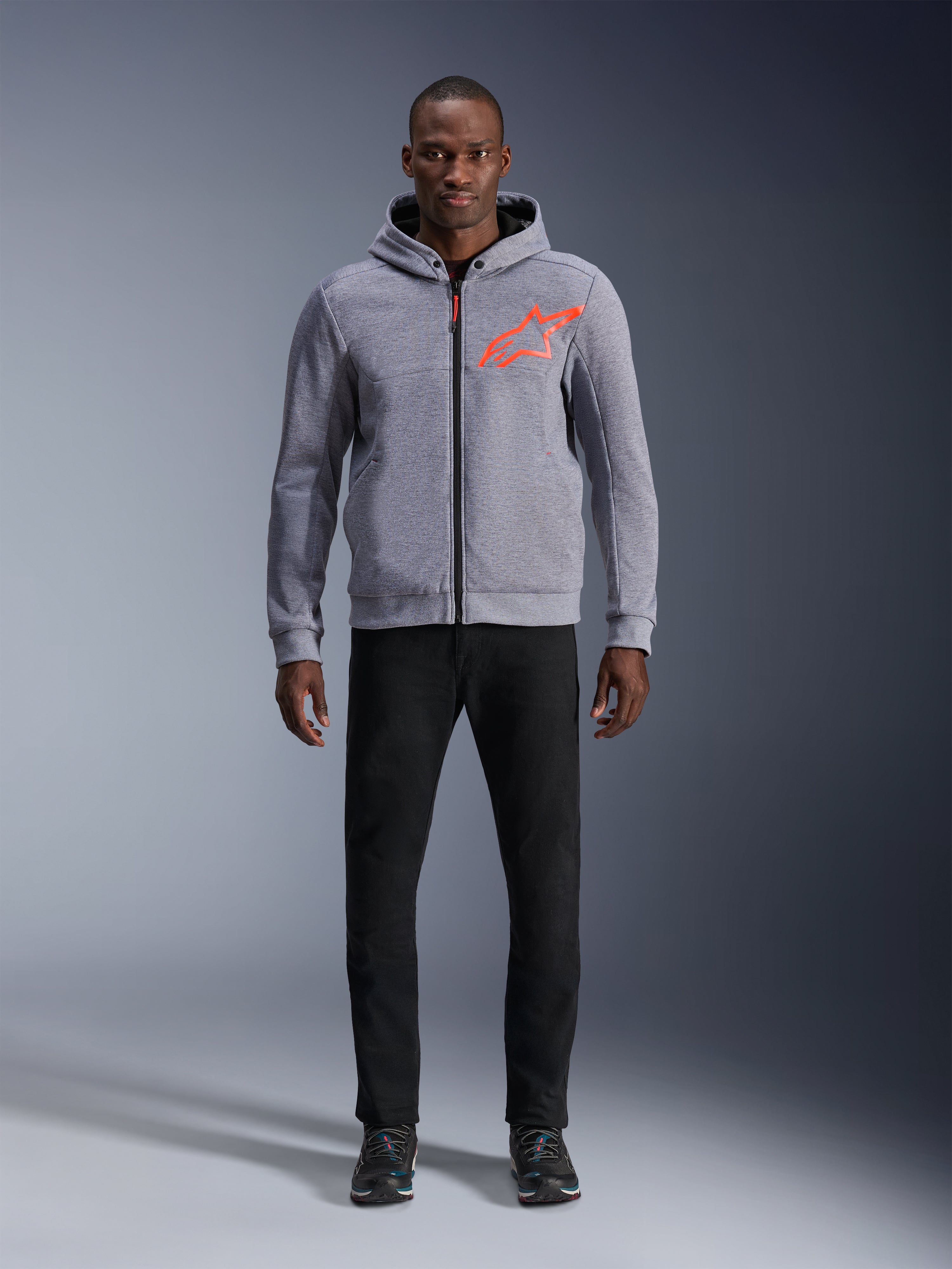 Modello che indossa Alpinestars Chrome V2 Sport Hoodie in Grigio Melange Rosso Fluorescente, felpa con cappuccio in pile tecnico grigio chiaro con logo a stella rosso arancio acceso sul petto, abbinata a pantaloni neri e sneakers scure, stile motociclistico urbano casual