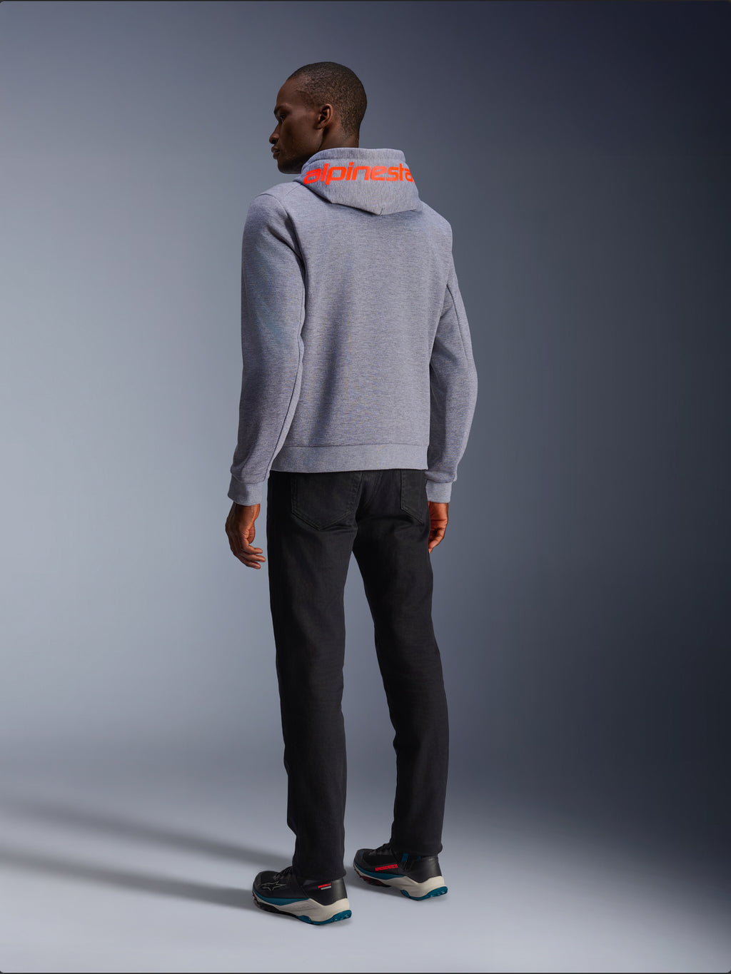 Alpinestars Chrome V2 Sport Hoodie in grigio melange, vista posteriore con grande logo Alpinestars rosso fluorescente sul cappuccio, indossata con pantaloni neri e scarpe da moto scure, abbigliamento moto casual urbano
