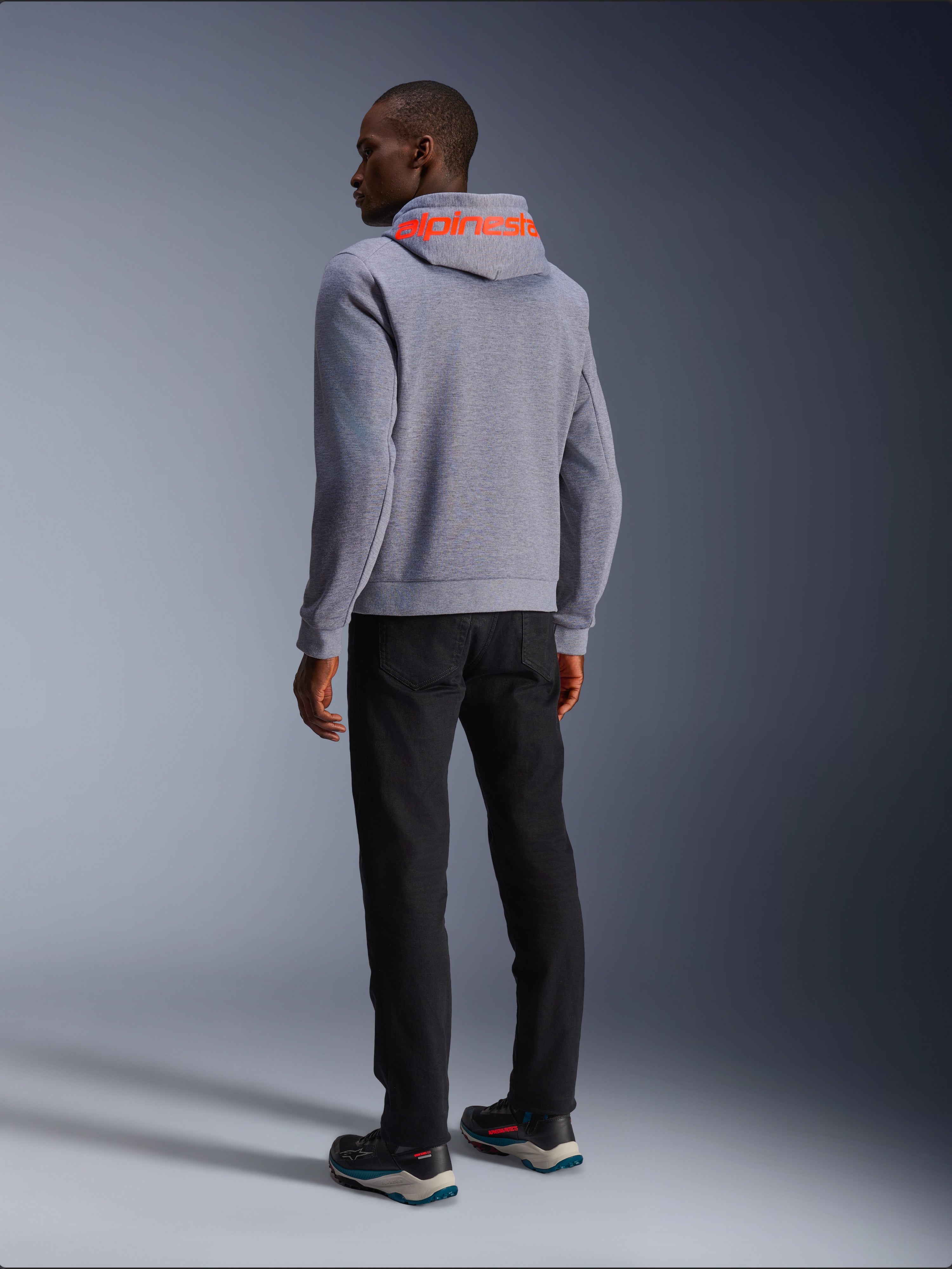 Alpinestars Chrome V2 Sport Hoodie in grigio melange, vista posteriore con grande logo Alpinestars rosso fluorescente sul cappuccio, indossata con pantaloni neri e scarpe da moto scure, abbigliamento moto casual urbano