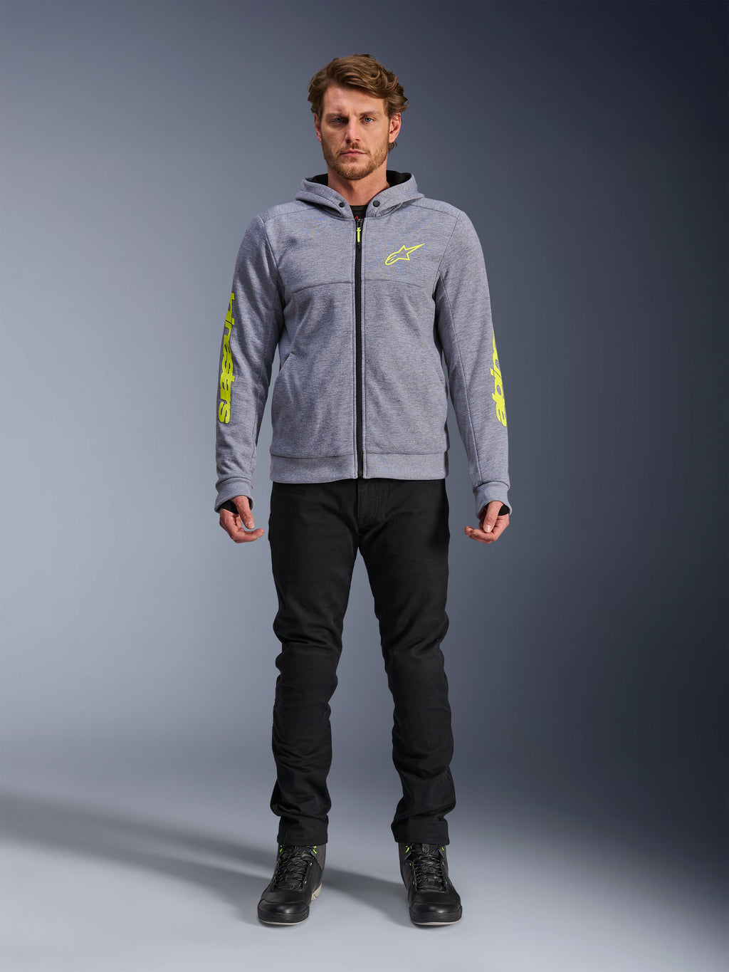 Alpinestars Chrome V2 Sport Hoodie, grigio mélange con accenti giallo fluorescente, abbigliamento moto urbano, indossata con pantaloni neri e scarpe da moto nere, pile tecnico con branding giallo sulle maniche e sul petto, stile street casual per la guida
