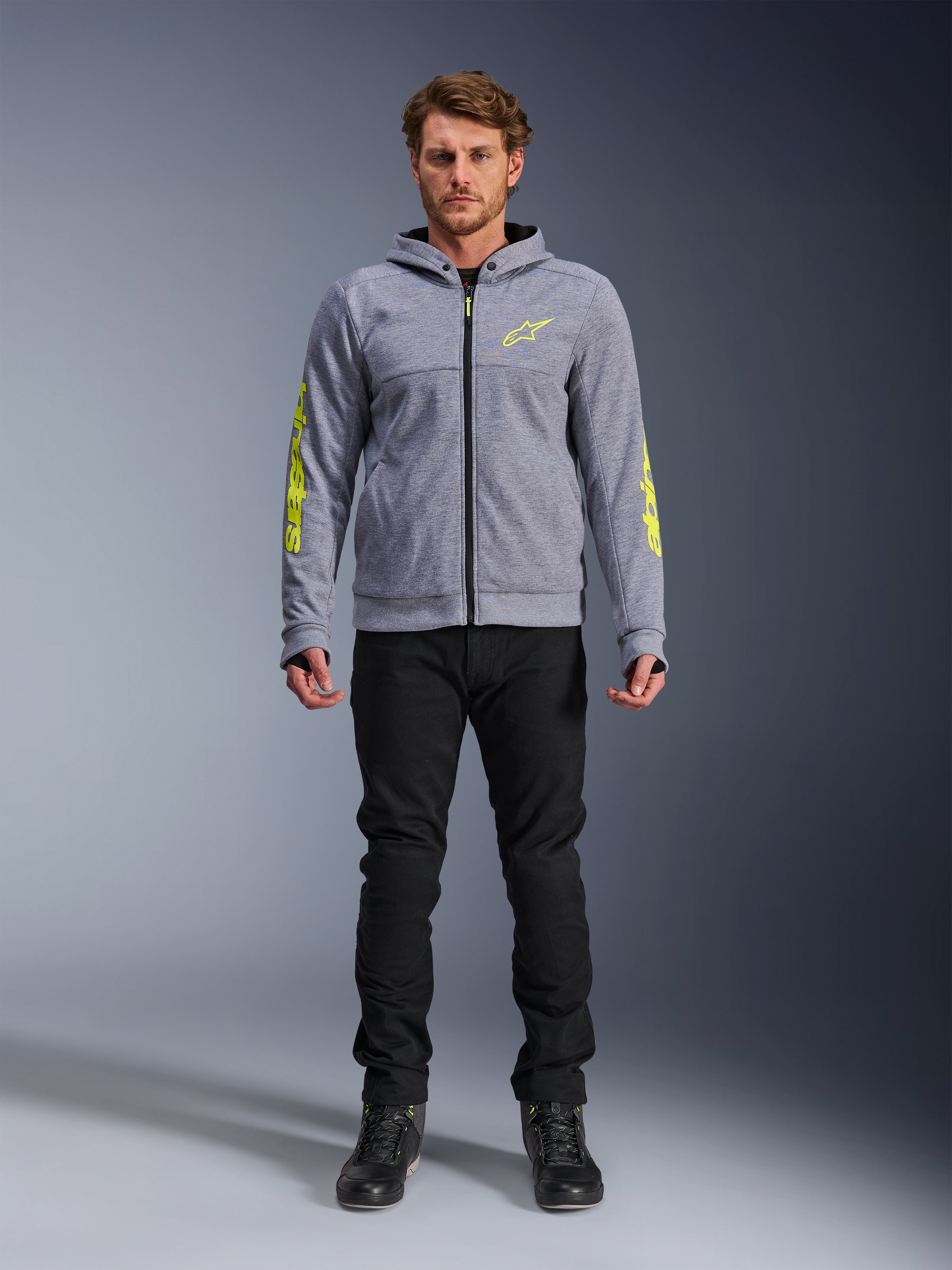 Alpinestars Chrome V2 Sport Hoodie, grigio mélange con accenti giallo fluorescente, abbigliamento moto urbano, indossata con pantaloni neri e scarpe da moto nere, pile tecnico con branding giallo sulle maniche e sul petto, stile street casual per la guida