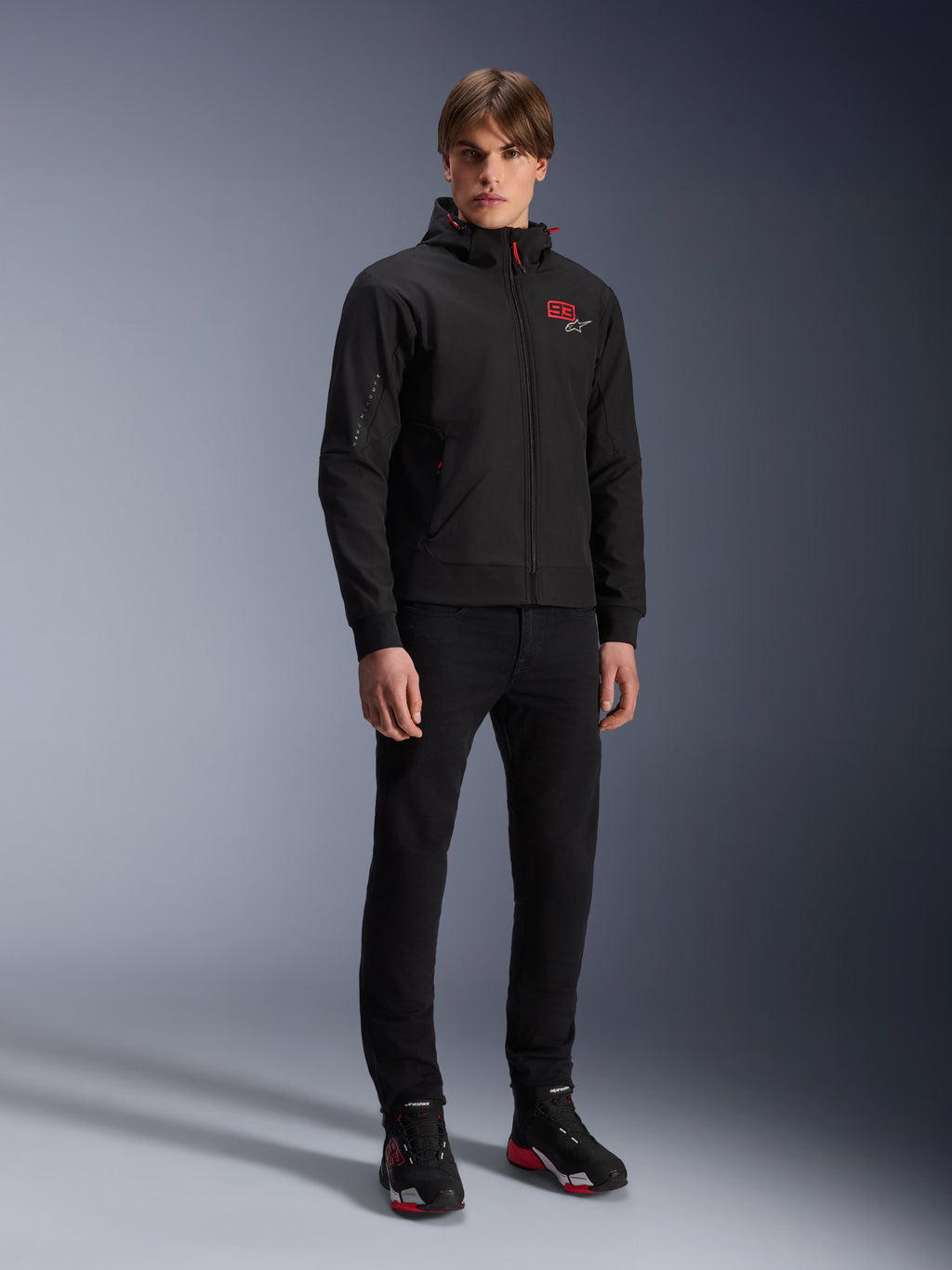 Alpinestars MM93 Radium Tech Hoodie, giacca da moto urban, nero con accenti rosso brillante, indossata da un modello con pantaloni neri e scarpe da moto, con logo '93' rosso sul petto e marchio Marc Marquez sulla manica, streetwear tecnico casual per la guida urbana