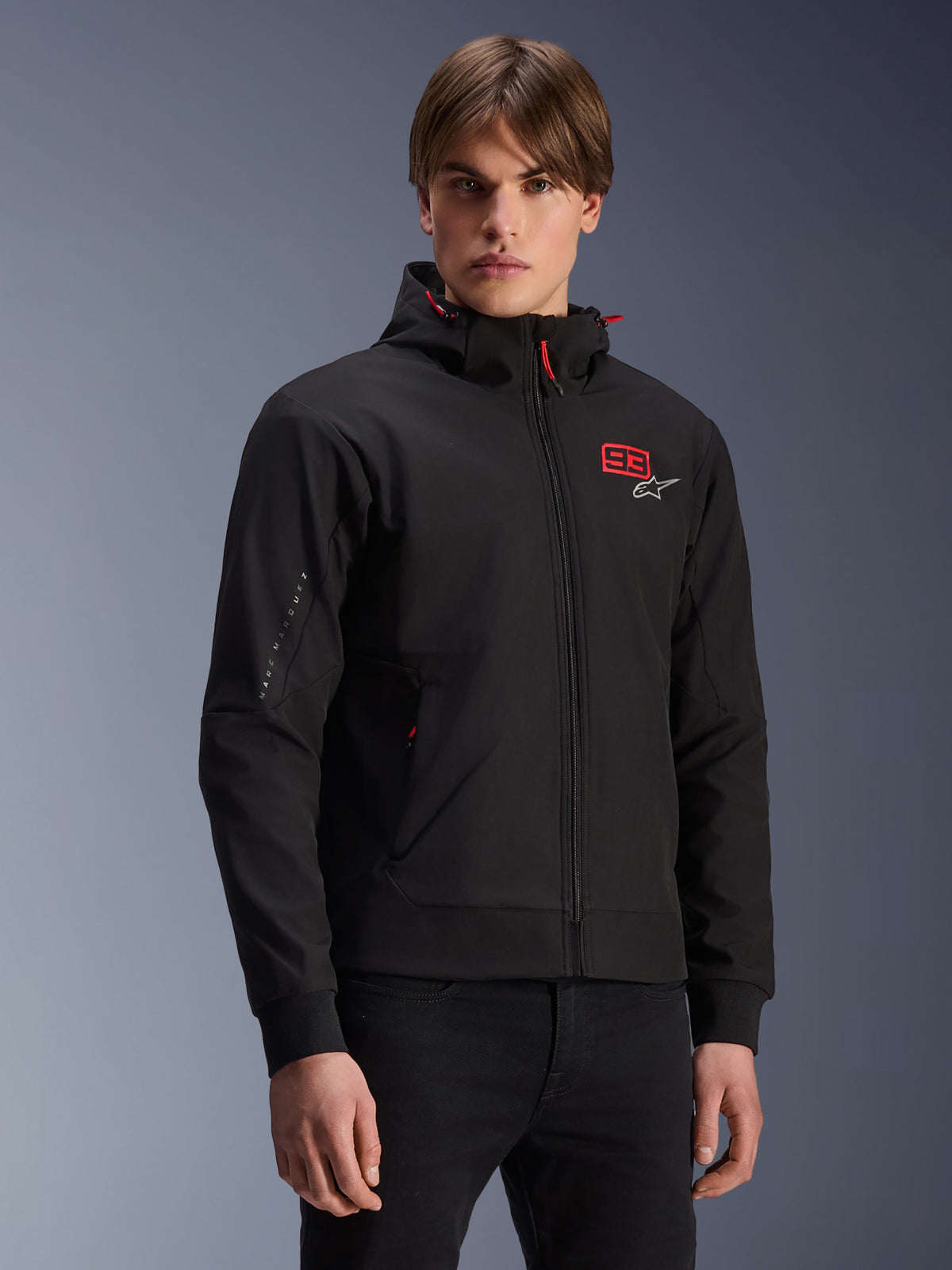 Alpinestars MM93 Radium Tech Hoodie, giacca da moto urbana nera con zip, numero 93 rosso e logo Alpinestars bianco sul petto, indossata da un modello con pantaloni neri, dotata di cappuccio rimovibile con cordini rossi e branding sulla manica, design casual ispirato al motorsport