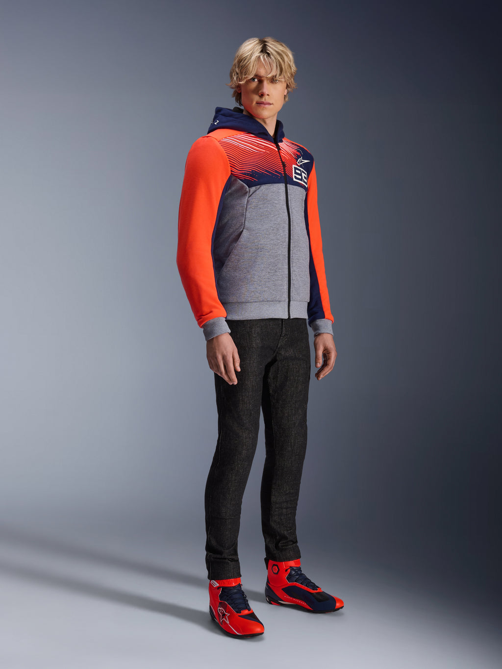 Alpinestars MM93 Chrome V2 Sport Hoodie, felpa da moto urbana, corpo grigio melange con petto blu navy e maniche rosso fluo acceso, con logo 93 sul petto, indossata da un modello con pantaloni neri slim-fit e scarpe da moto alte coordinate rosse e blu, vestibilità atletica moderna per la guida urbana