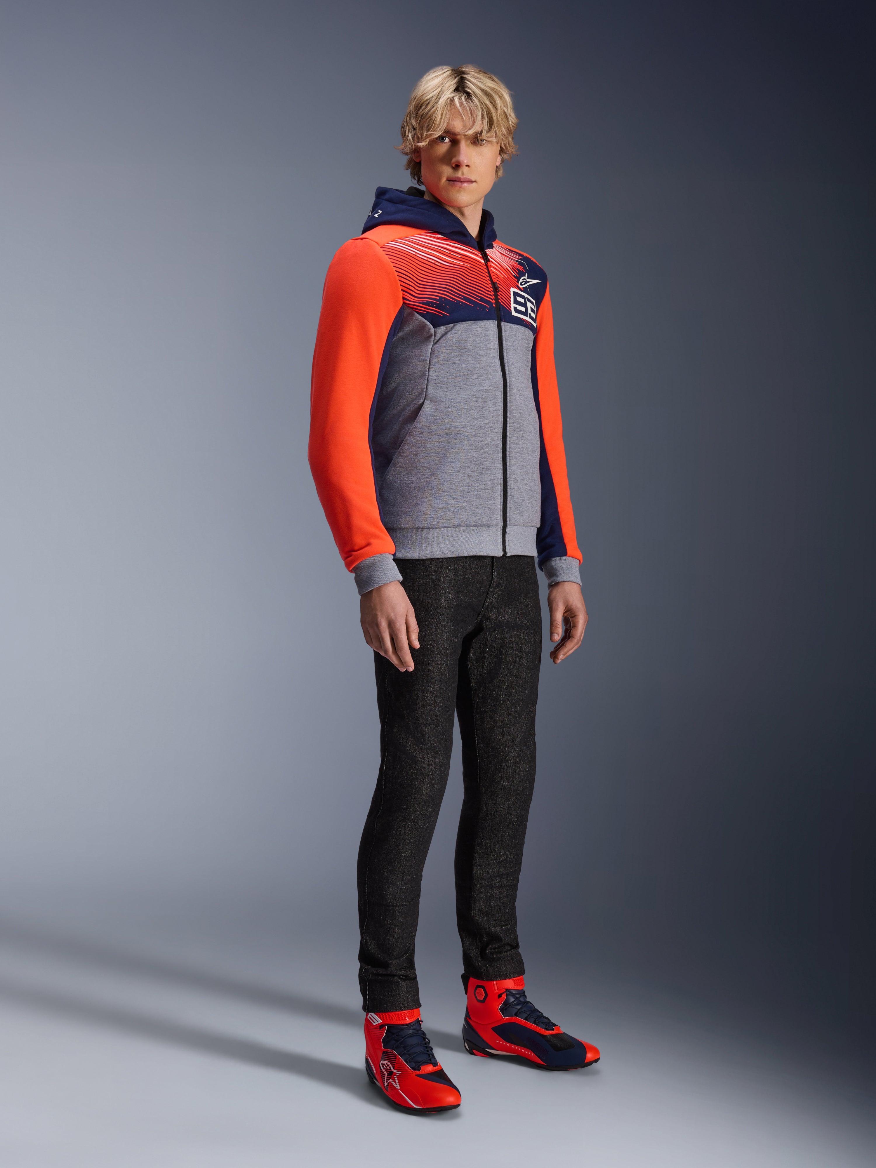 Alpinestars MM93 Chrome V2 Sport Hoodie, felpa da moto urbana, corpo grigio melange con petto blu navy e maniche rosso fluo acceso, con logo 93 sul petto, indossata da un modello con pantaloni neri slim-fit e scarpe da moto alte coordinate rosse e blu, vestibilità atletica moderna per la guida urbana