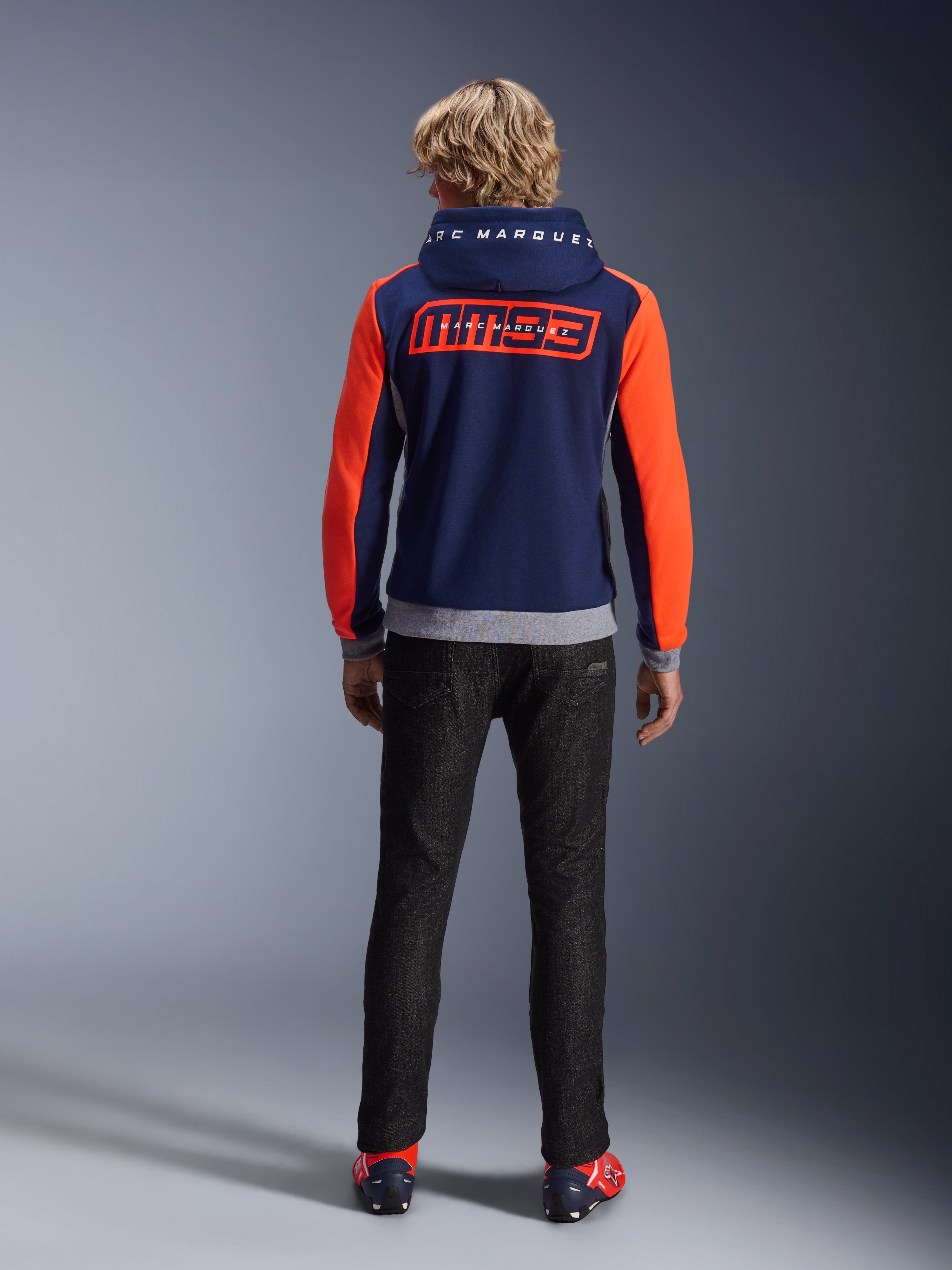 Modello visto di spalle che indossa la felpa Alpinestars MM93 Chrome V2 Sport Hoodie, blu navy con maniche rosso fluo e dettagli grigio melange, grandi loghi MM93 e Marc Marquez sulla schiena e sul cappuccio, felpa da moto casual abbinata a pantaloni neri