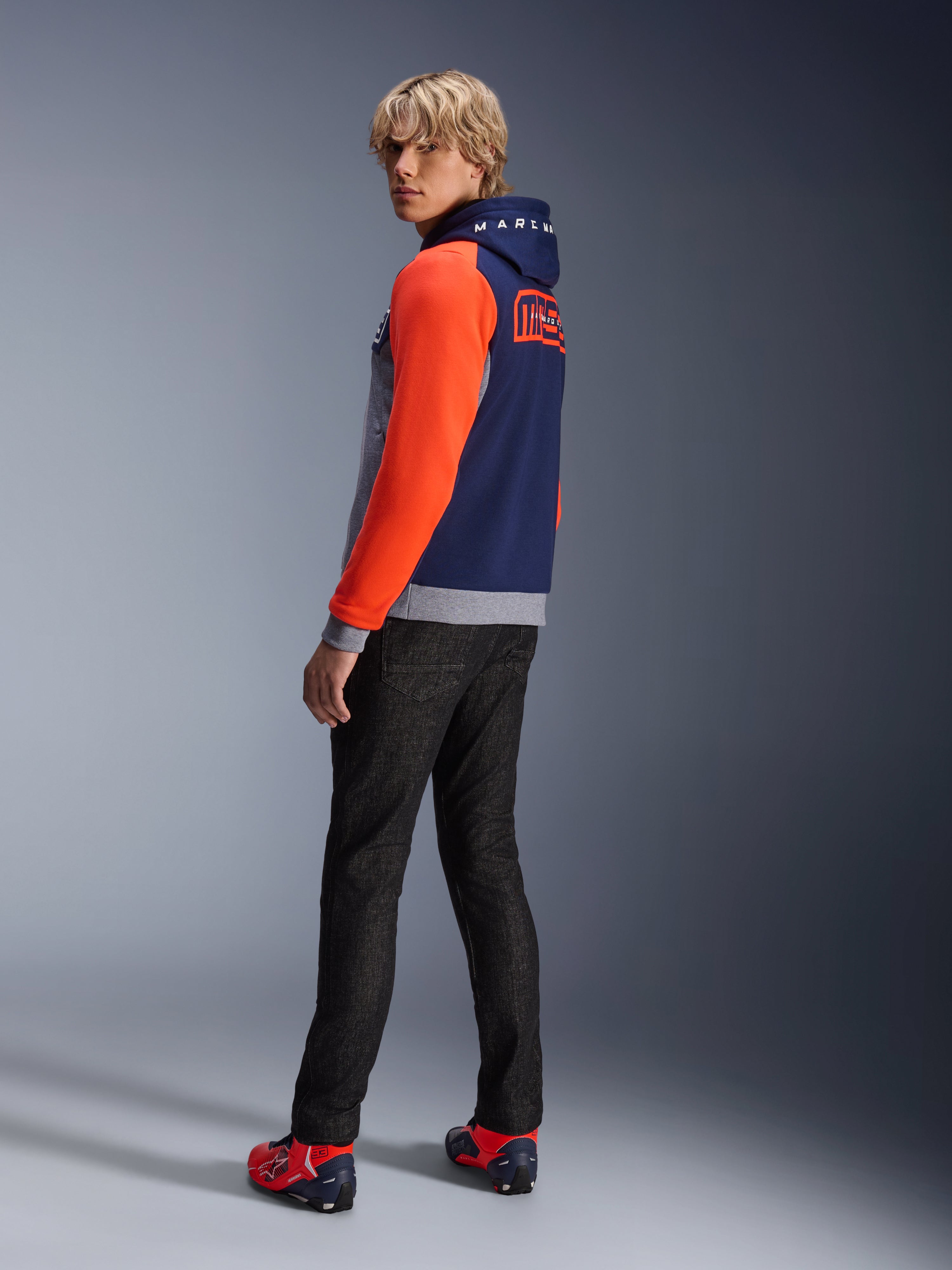 Alpinestars MM93 Chrome V2 Sport Hoodie, Blue Navy Grigio Melange Rosso Fluoro, modello che indossa la felpa con maniche rosso acceso e branding MM93 Marc Marquez sul retro, abbinata a jeans scuri e scarpe da moto rosse, abbigliamento moto urban casual