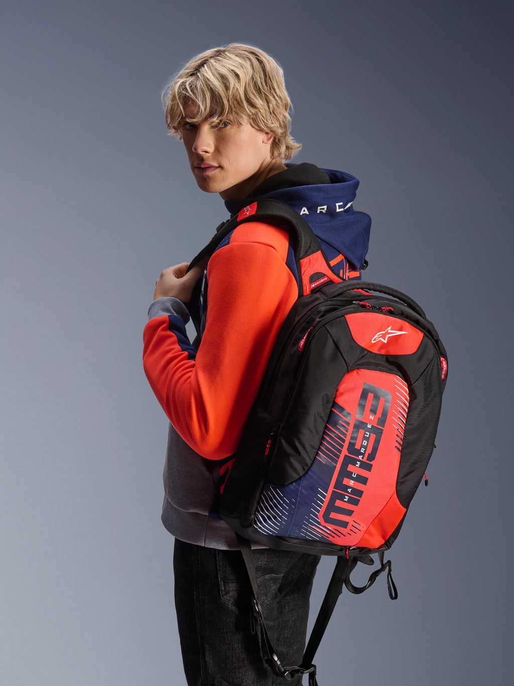 Modello che indossa la felpa con cappuccio Alpinestars MM93 Chrome V2 Sport in blu navy, grigio melange e rosso fluo, abbinata a uno zaino MM93 coordinato, abbigliamento da moto urbano casual con silhouette atletica moderna.