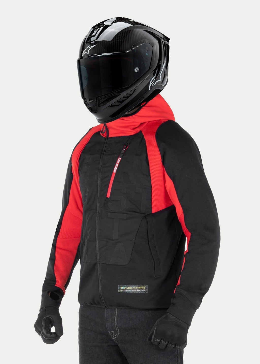 Giacca con cappuccio Alpinestars MO.ST.EQ Hybrid, felpa da moto urbana, nero e rosso, con cerniera anteriore angolata e pannelli a coste sulle spalle, indossata con casco integrale nero e guanti neri, stile streetwear contemporaneo