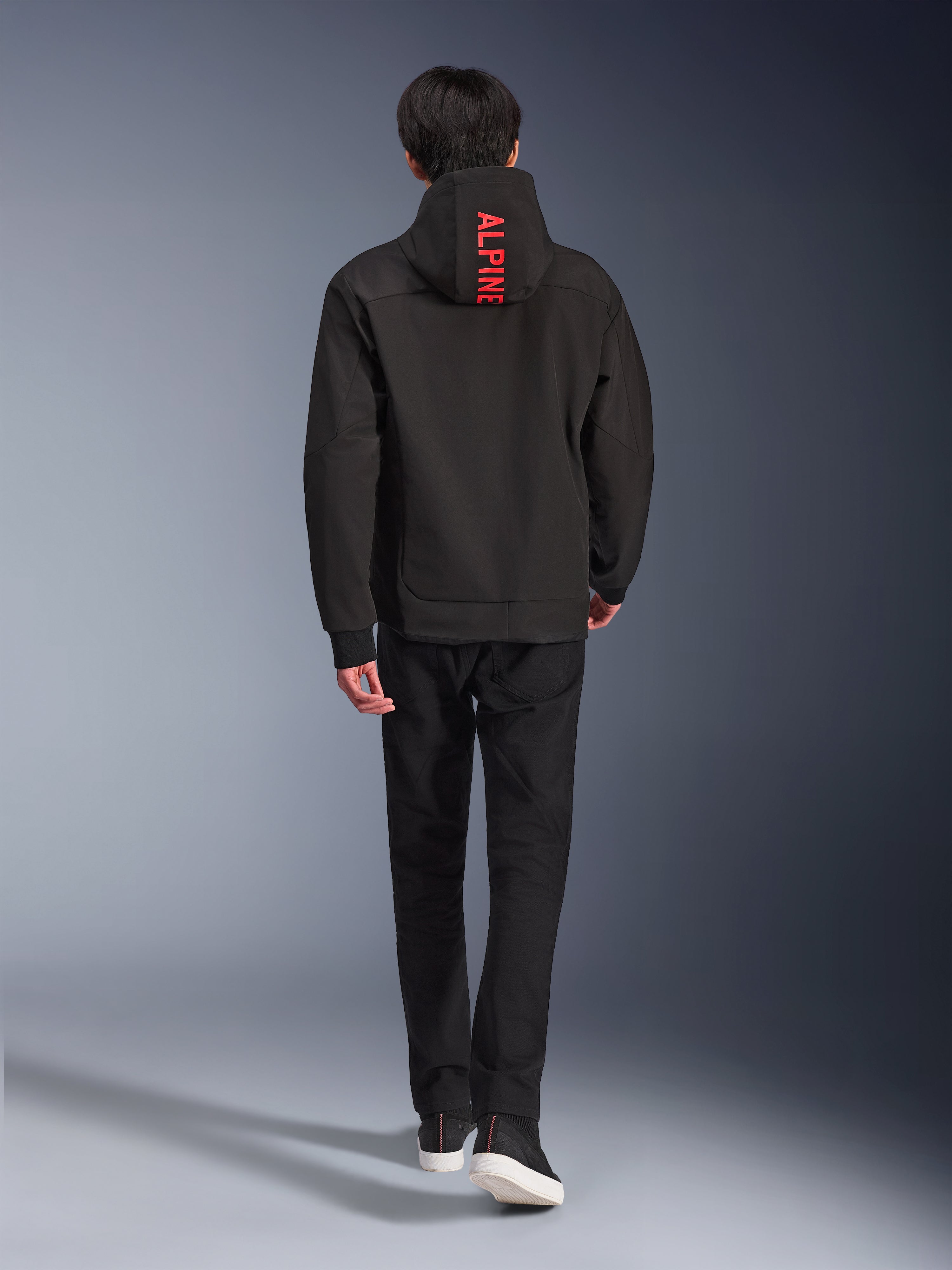Alpinestars Radium Tech Hoodie, giacca da moto urbana, nera, vista posteriore con scritta 'ALPINE' in rosso acceso sul cappuccio, indossata con pantaloni neri e sneakers, guscio tecnico elegante per il pendolarismo.
