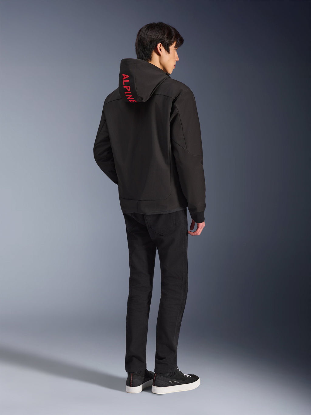 Alpinestars Radium Tech Hoodie, nera con marchio Alpinestars rosso sul cappuccio, vista posteriore su un modello maschile, indossata con pantaloni neri e sneakers, abbigliamento tecnico per guida urbana