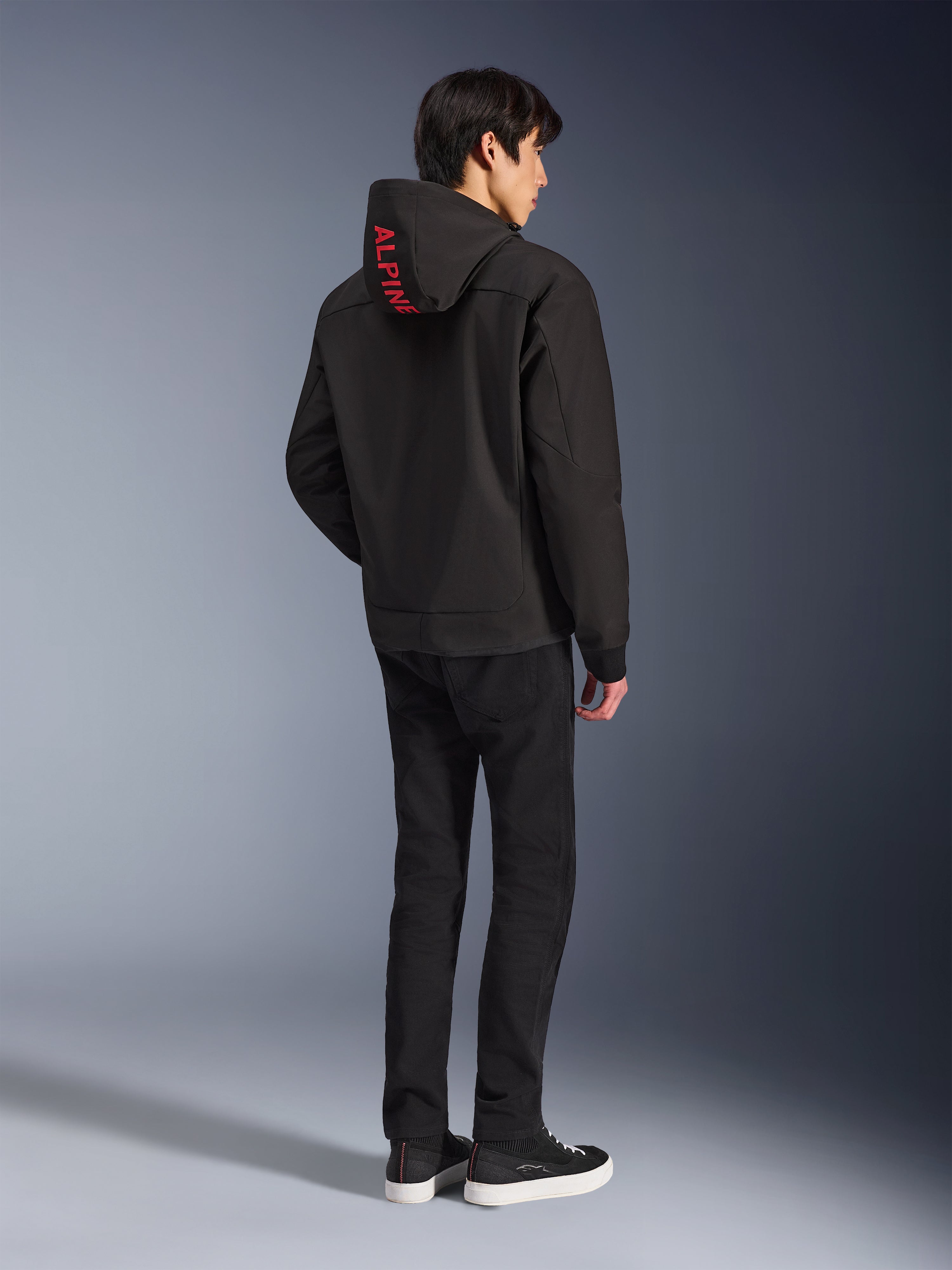 Alpinestars Radium Tech Hoodie, nera con marchio Alpinestars rosso sul cappuccio, vista posteriore su un modello maschile, indossata con pantaloni neri e sneakers, abbigliamento tecnico per guida urbana